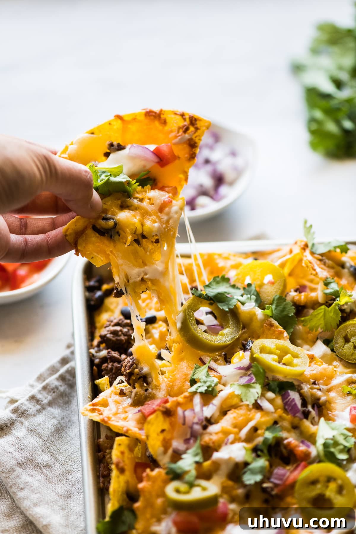 Loaded Sheet Pan Nachos 3 A tortilla chip topped with melted cheese being pulled off a pan of sheet pan nachos, showcasing a perfect cheese pull.