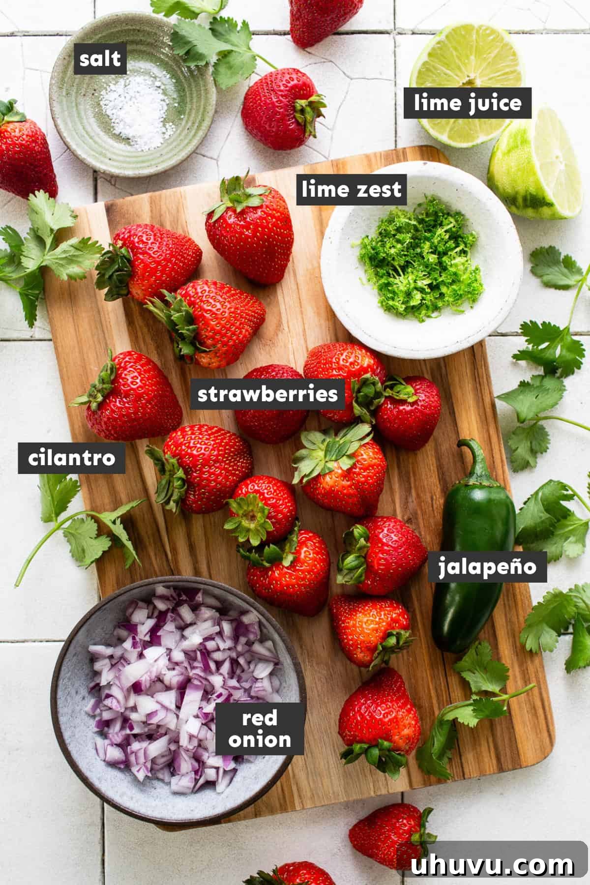 A vibrant selection of fresh ingredients for strawberry salsa, including bright red strawberries, a green jalapeño, a purple red onion, fresh cilantro, and two limes, arranged on a rustic wooden table.