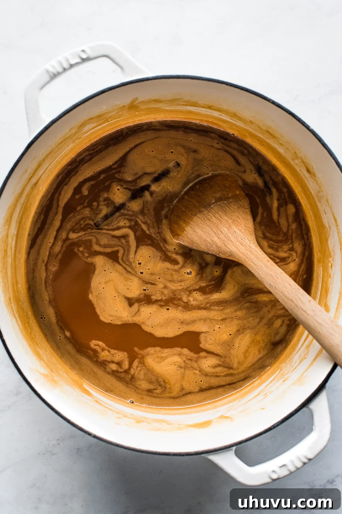 A wooden spoon stirring cajeta in a dutch oven. The mixture is simmering and turning golden.