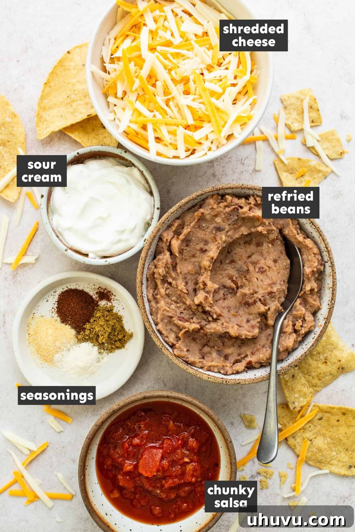 Savory Fiesta Bean Dip 3 An array of fresh ingredients for bean dip, neatly arranged on a rustic wooden table, showcasing refried beans, salsa, sour cream, various spices, and a block of cheese, ready for preparation.