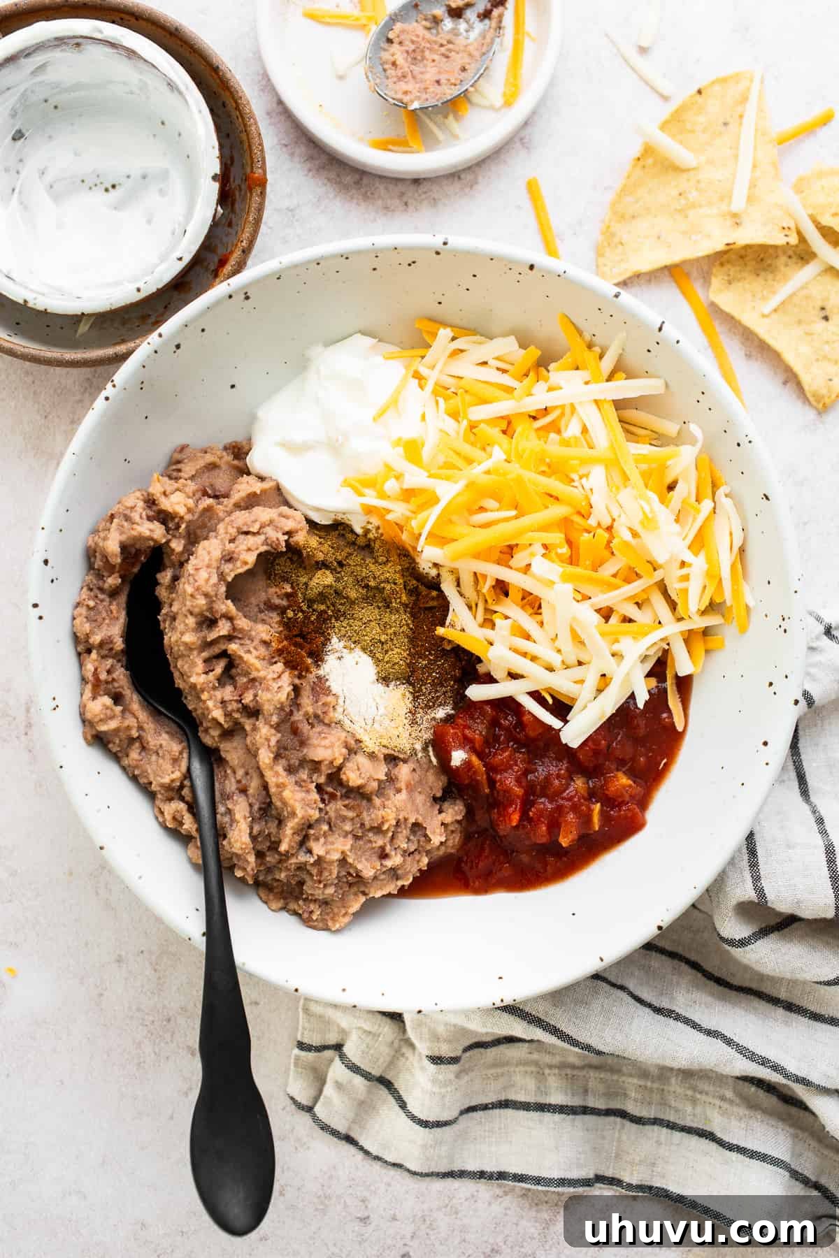 Savory Fiesta Bean Dip 4 A large mixing bowl containing refried beans, chunky salsa, a portion of shredded cheese, creamy sour cream, and a medley of seasonings, all ready to be combined into a flavorful bean dip base.