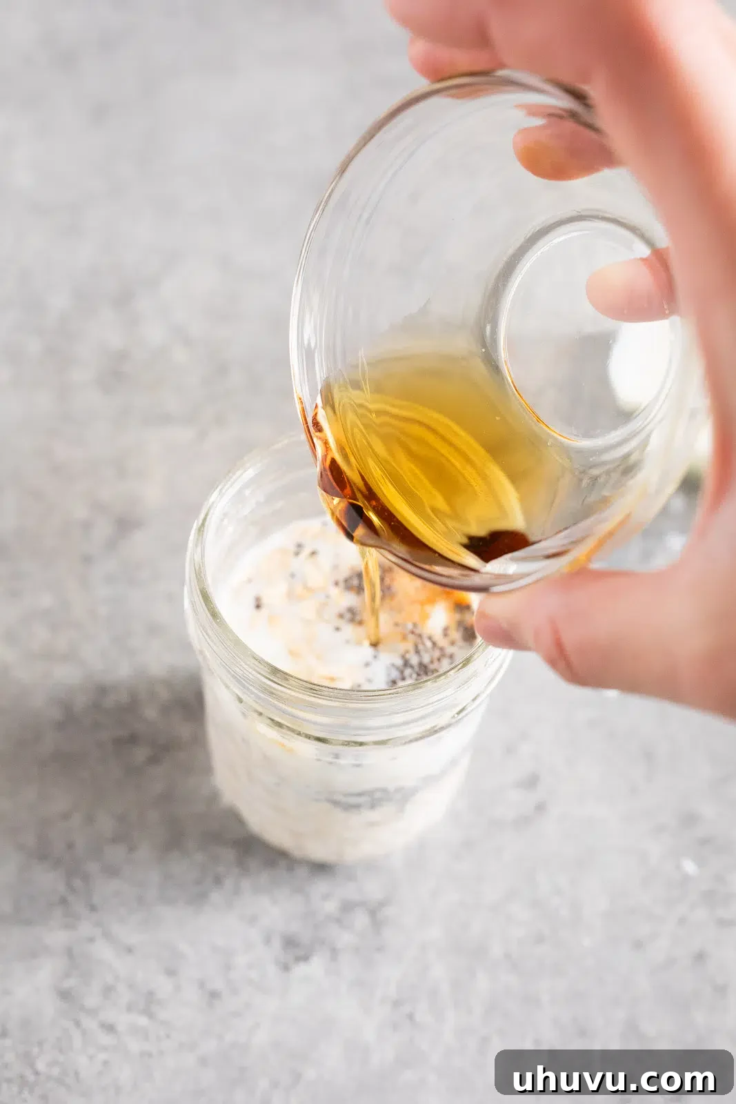A hand adding rolled oats into a glass jar, followed by milk and honey, demonstrating the initial step of preparing overnight oats.