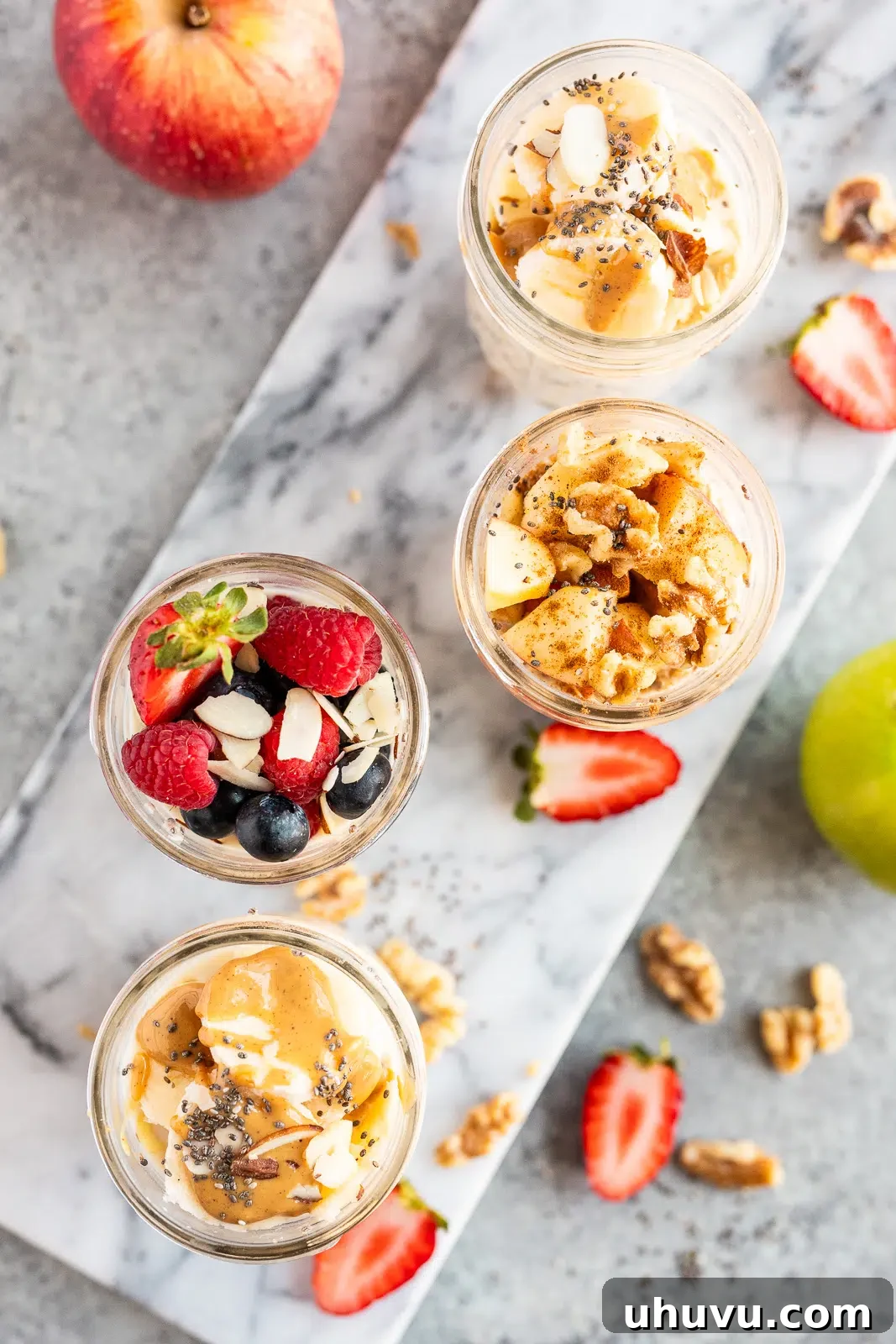 An overhead shot of four glass jars filled with overnight oats, each topped with different combinations of fresh berries, apple slices, cinnamon, walnuts, and peanut butter, illustrating diverse meal prep options.