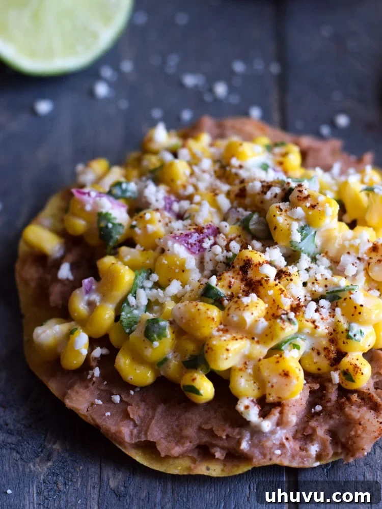 Zesty Elote Tostadas 4 A single Mexican Street Corn Tostada, beautifully topped with a layer of creamy refried beans, a generous scoop of vibrant Mexican street corn, and sprinkled with extra Cotija cheese, presented on a wooden surface.