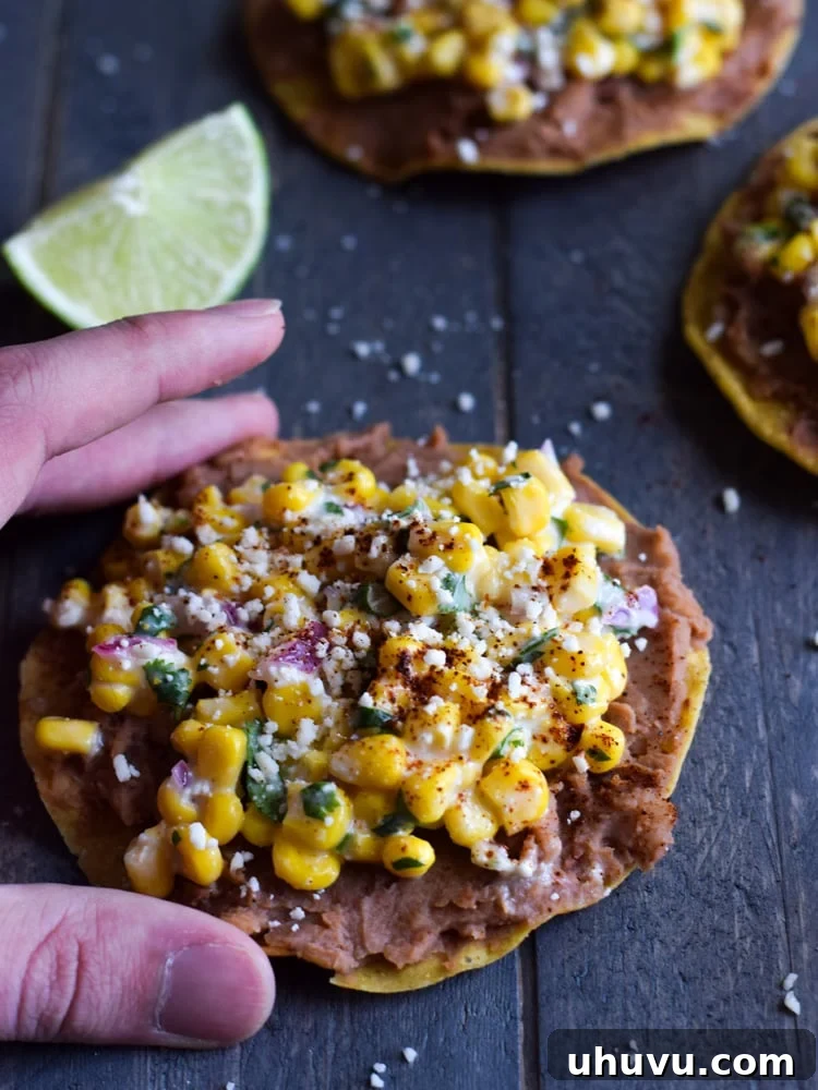 Zesty Elote Tostadas 6 A hand reaching for a crisp Mexican Street Corn Tostada, showcasing its vibrant toppings of refried beans, street corn, and Cotija cheese. This image captures the deliciousness and ease of enjoying this quick, gluten-free, and vegetarian meal.