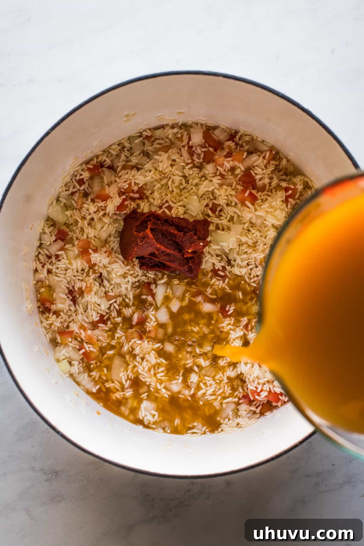 Savory broth being poured into a pot containing toasted rice, onion, and tomato to begin the simmering process for authentic Mexican rice.