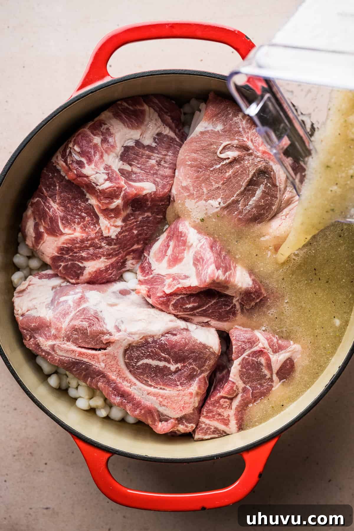 The smooth, blended broth mixture being carefully poured into a large pot containing chunks of pork shoulder and hominy, ready for simmering.