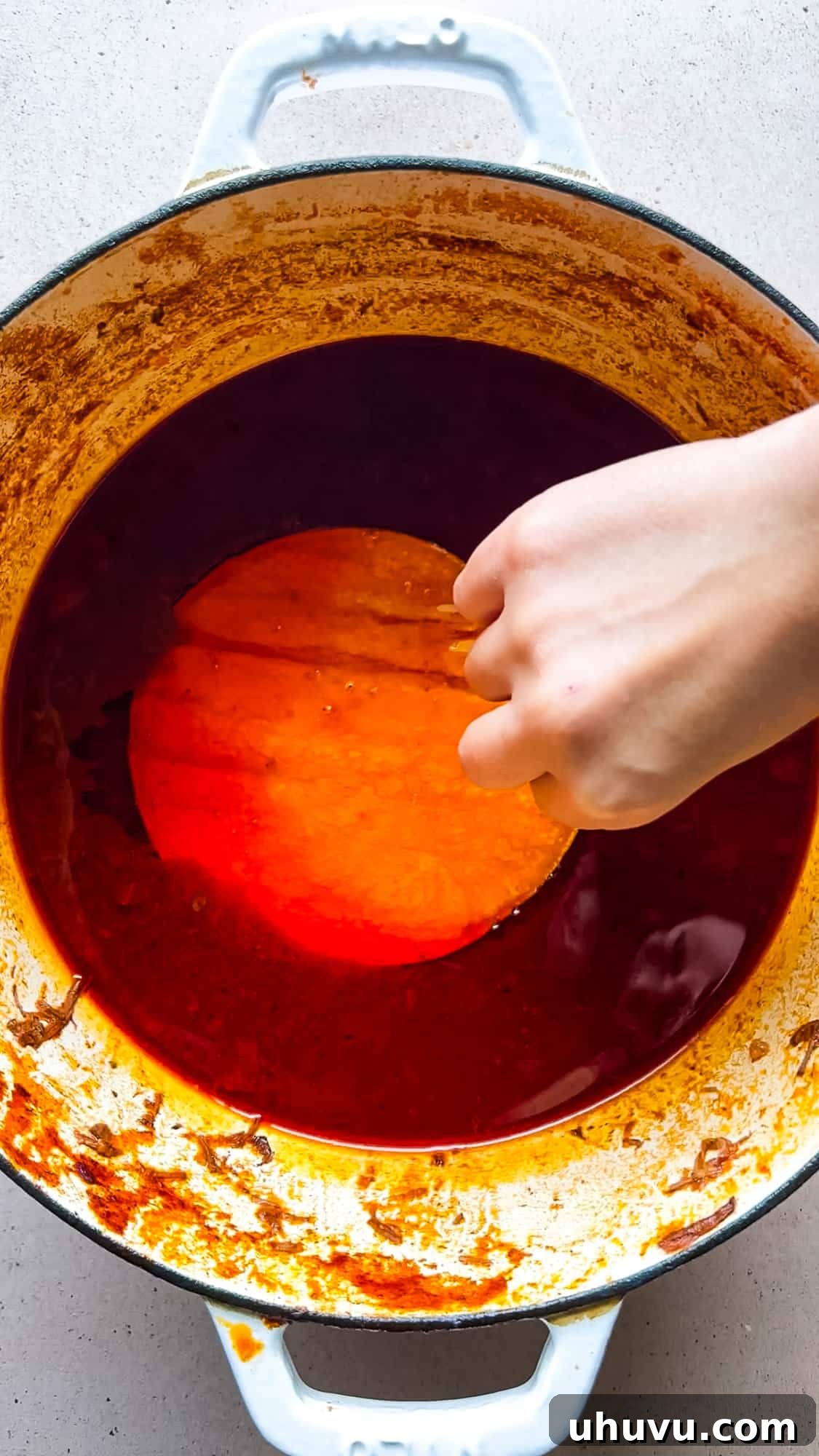 A corn tortilla being dipped into the top layer of fat of a pot of birria for making birria tacos.