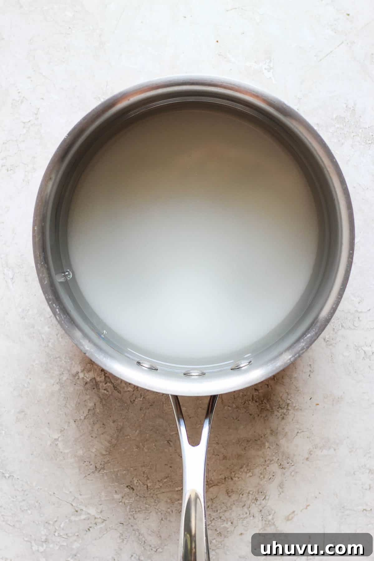 Close-up of clear water and white granulated sugar simmering together in a stainless steel saucepan on a stovetop, forming the base for homemade raspado syrup.