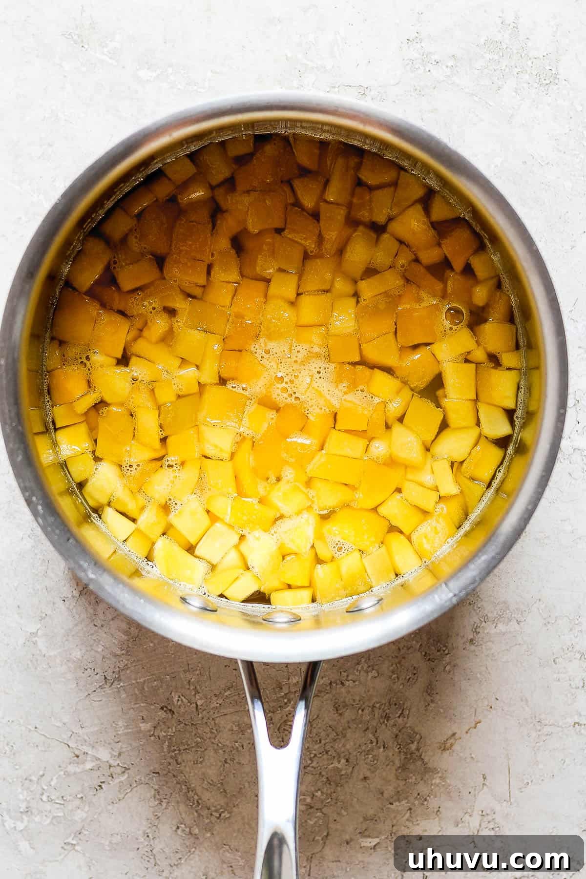 Diced mango pieces cooking in a saucepan with a clear simple syrup (water and sugar) on a stovetop, creating the base for delicious raspados syrup.