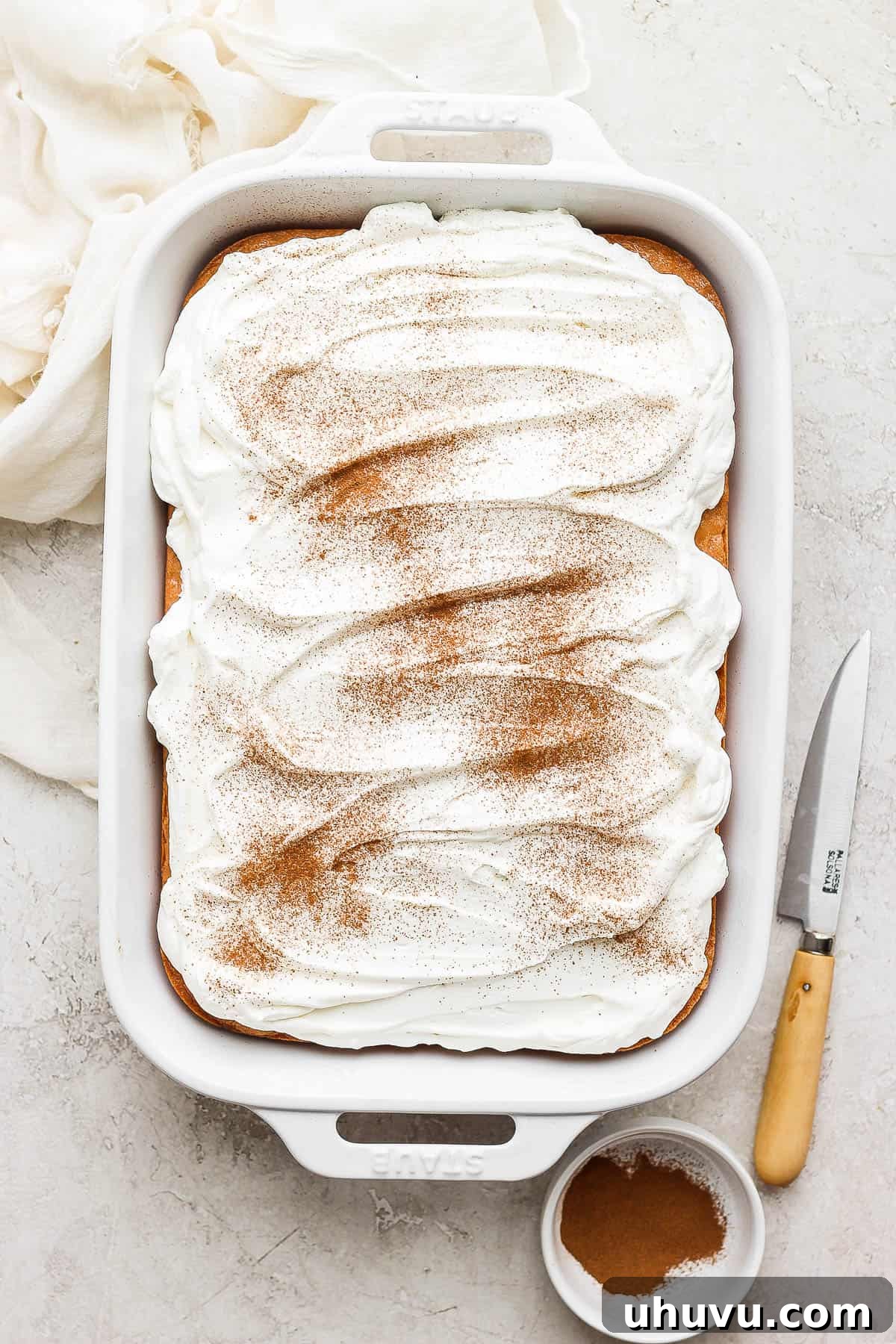 September's Kitchen Showdown 3 A perfectly baked Tres Leches Cake cooling in a baking pan, lavishly topped with airy whipped cream and a subtle sprinkle of ground cinnamon, ready for portioning.