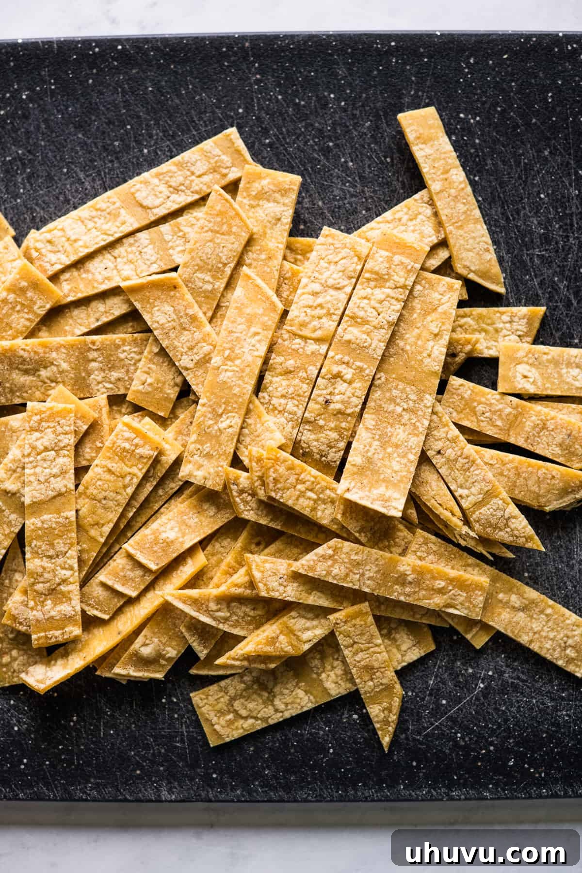 Tortilla Crisps 6 A pile of uncooked corn tortilla strips, neatly cut and arranged on a cutting board, awaiting the frying process.