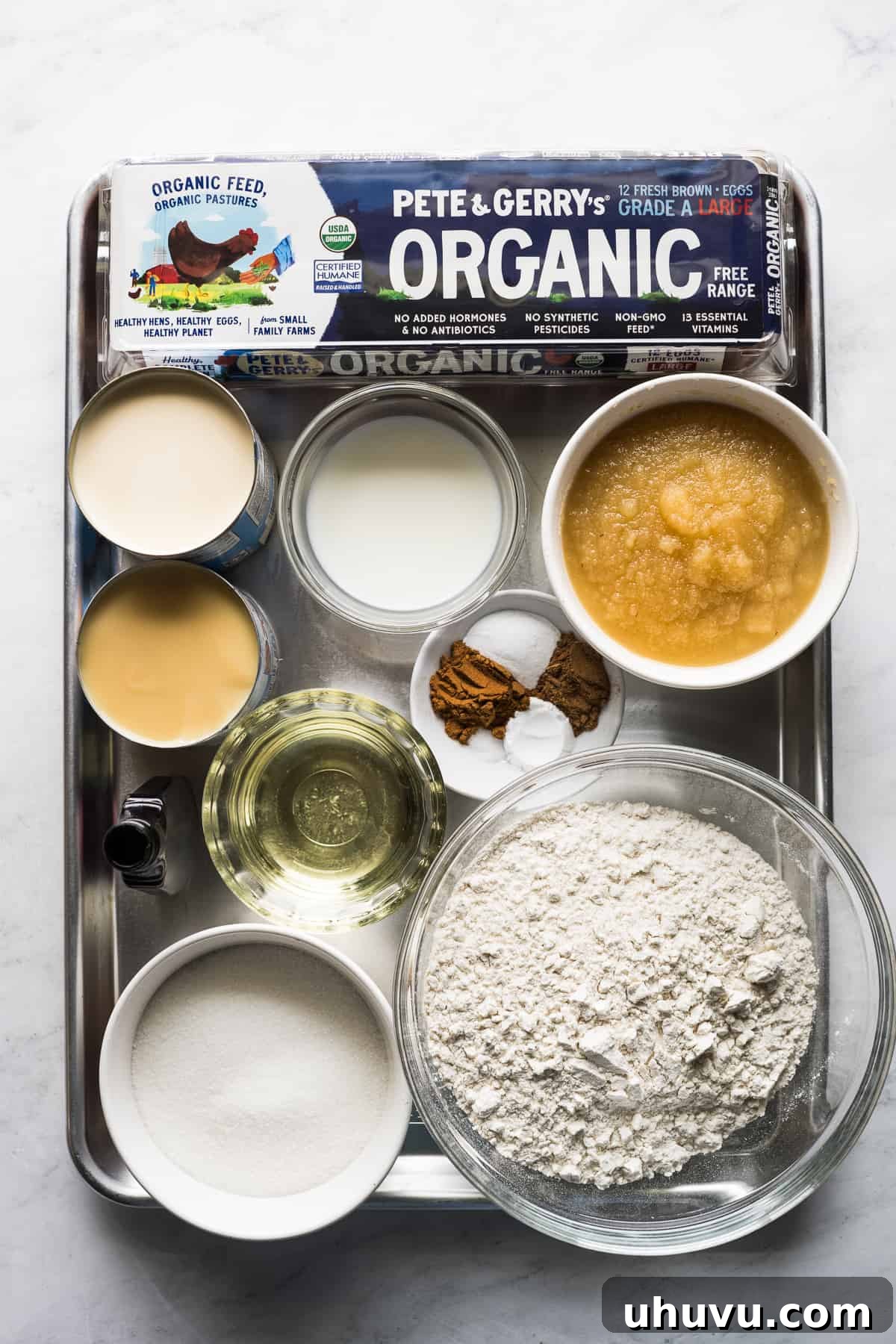 Spiced Apple Tres Leches Dream 3 Various ingredients for an apple tres leches cake laid out on a wooden table, including eggs, flour, applesauce, milks, and spices, highlighted as essential for this fall dessert recipe.