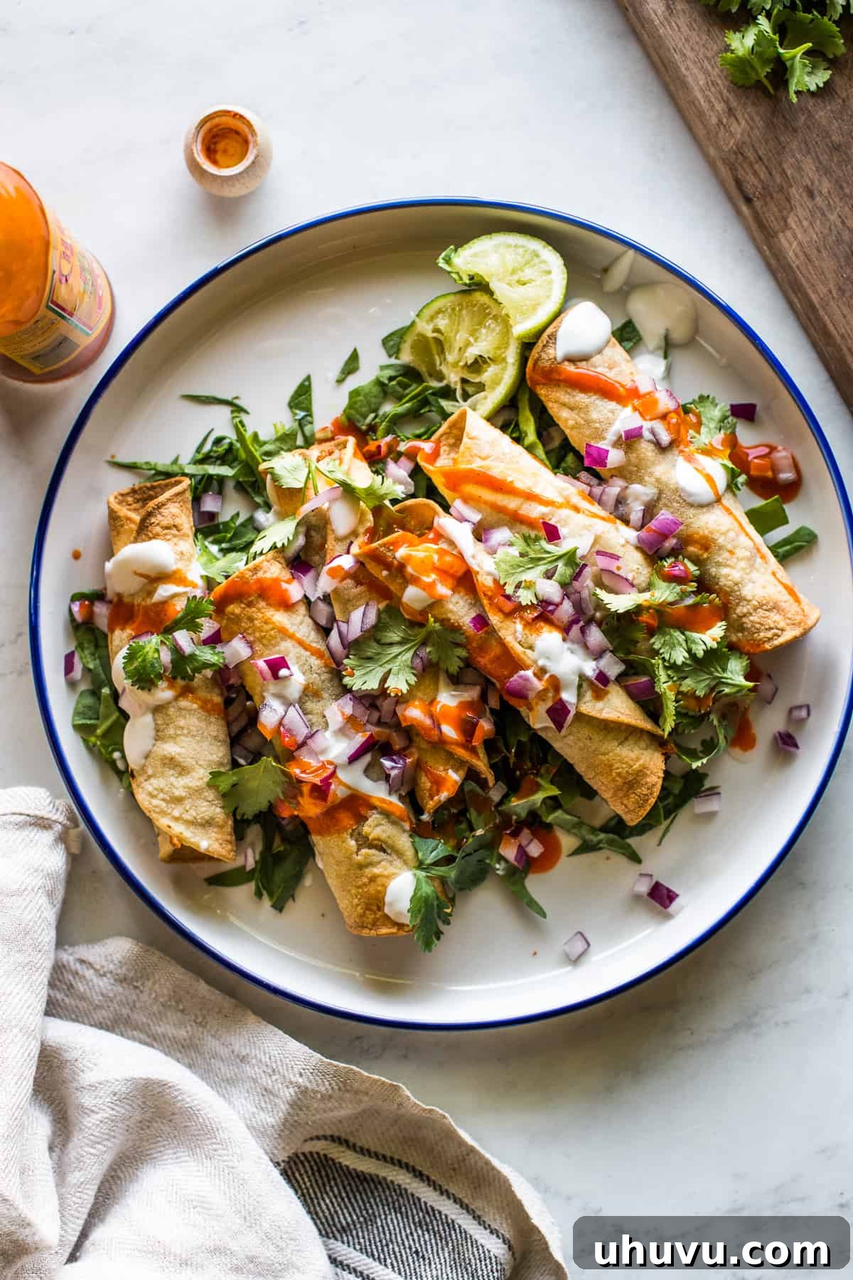 Air fryer chicken taquitos on a plate topped with Mexican crema, hot sauce, cilantro, and diced onion.