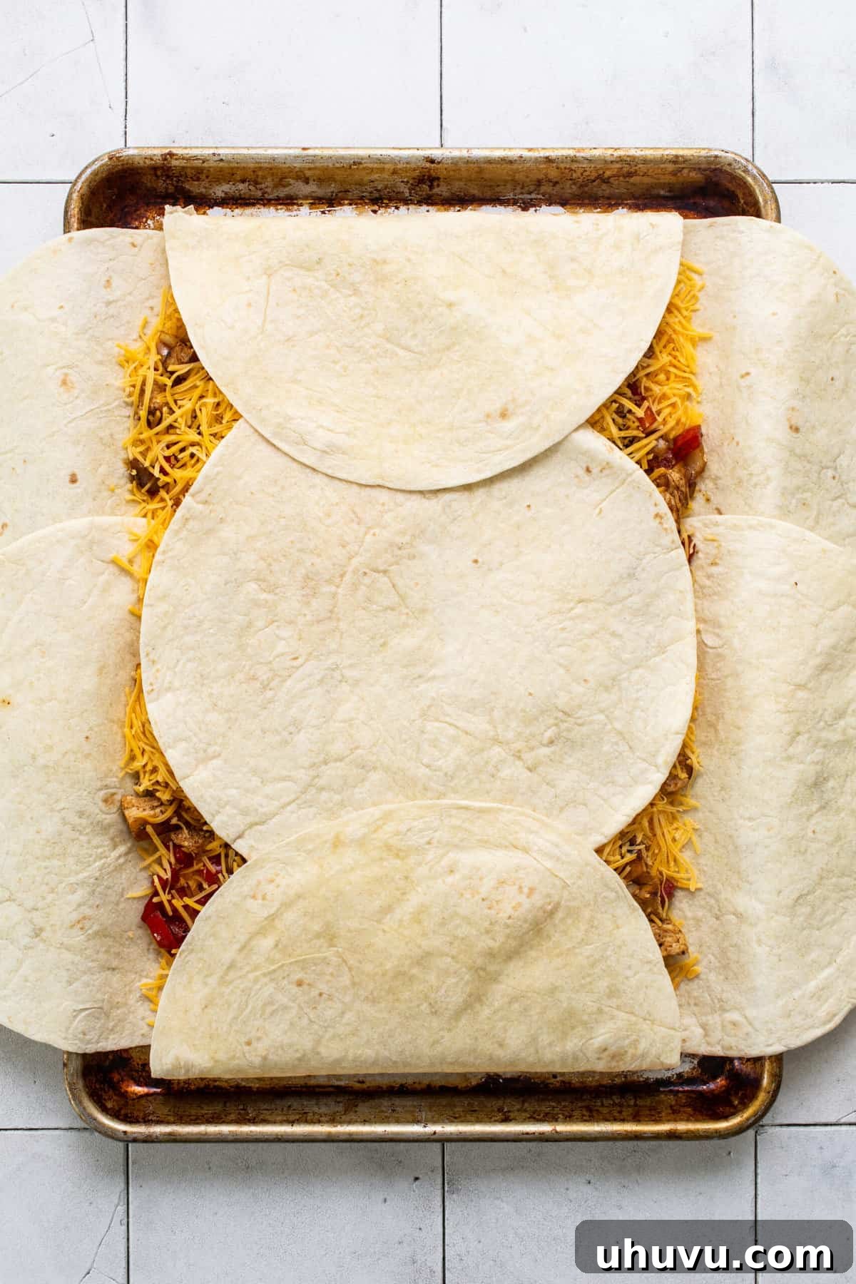 The outer flour tortillas of the sheet pan quesadilla are being folded inward over the filling, creating a large, enclosed quesadilla ready for its final top layer.