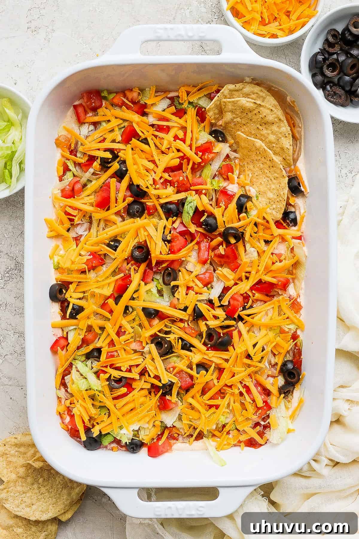 Ultimate Taco Party Dip 2 A vibrant taco dip served in a baking dish, generously topped with shredded cheese, diced tomatoes, and sliced black olives, accompanied by crispy tortilla chips, inviting guests to dig in.