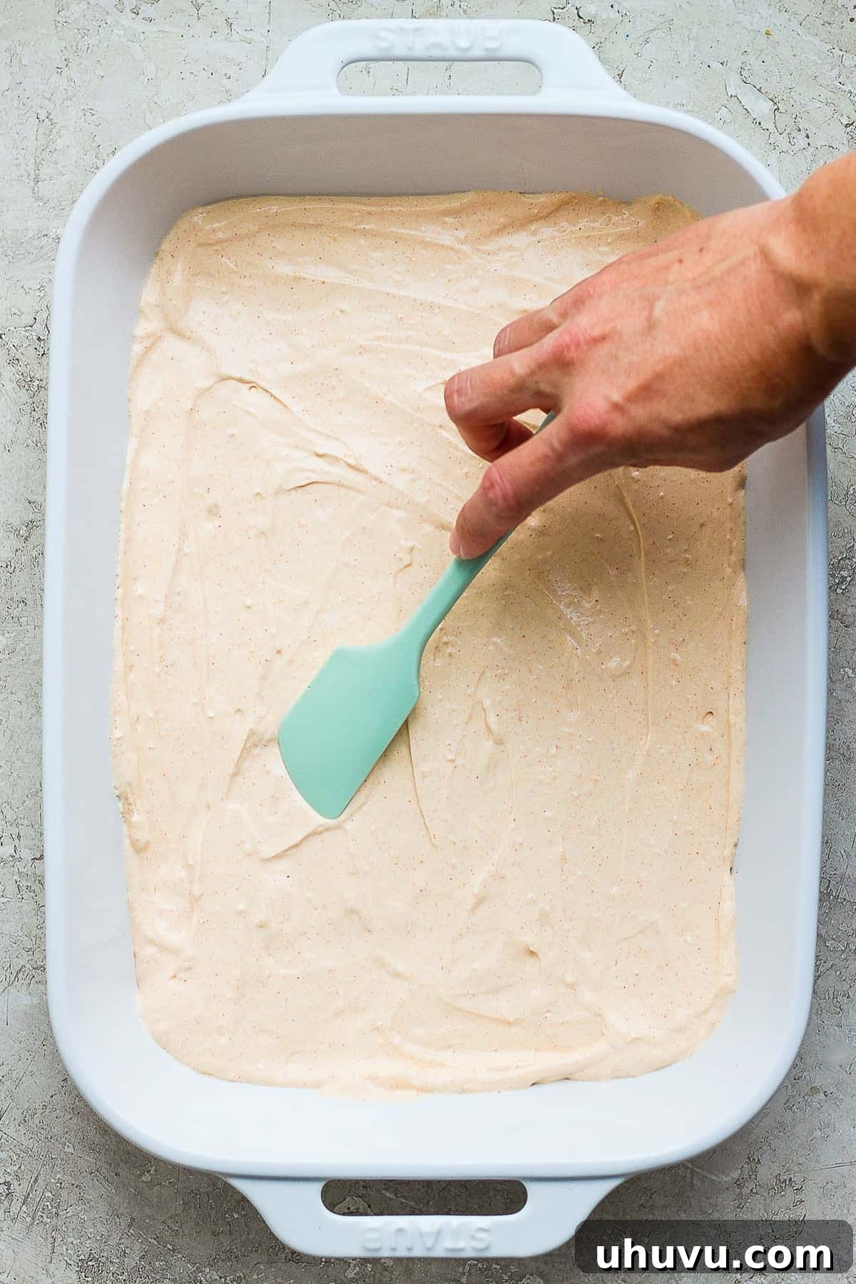 Ultimate Taco Party Dip 6 The smooth cream cheese and sour cream mixture of taco dip being carefully spread into a large 9x13-inch baking dish, forming an even base layer.