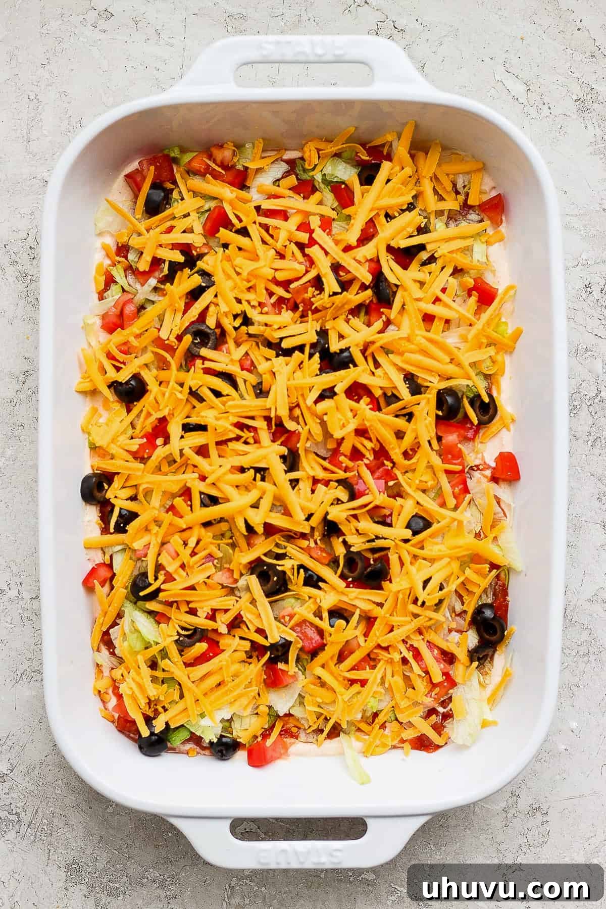 Ultimate Taco Party Dip 9 A completed taco dip, meticulously layered with all its toppings including cheese, tomatoes, and olives, beautifully presented in a casserole baking dish and ready to be served at a gathering.