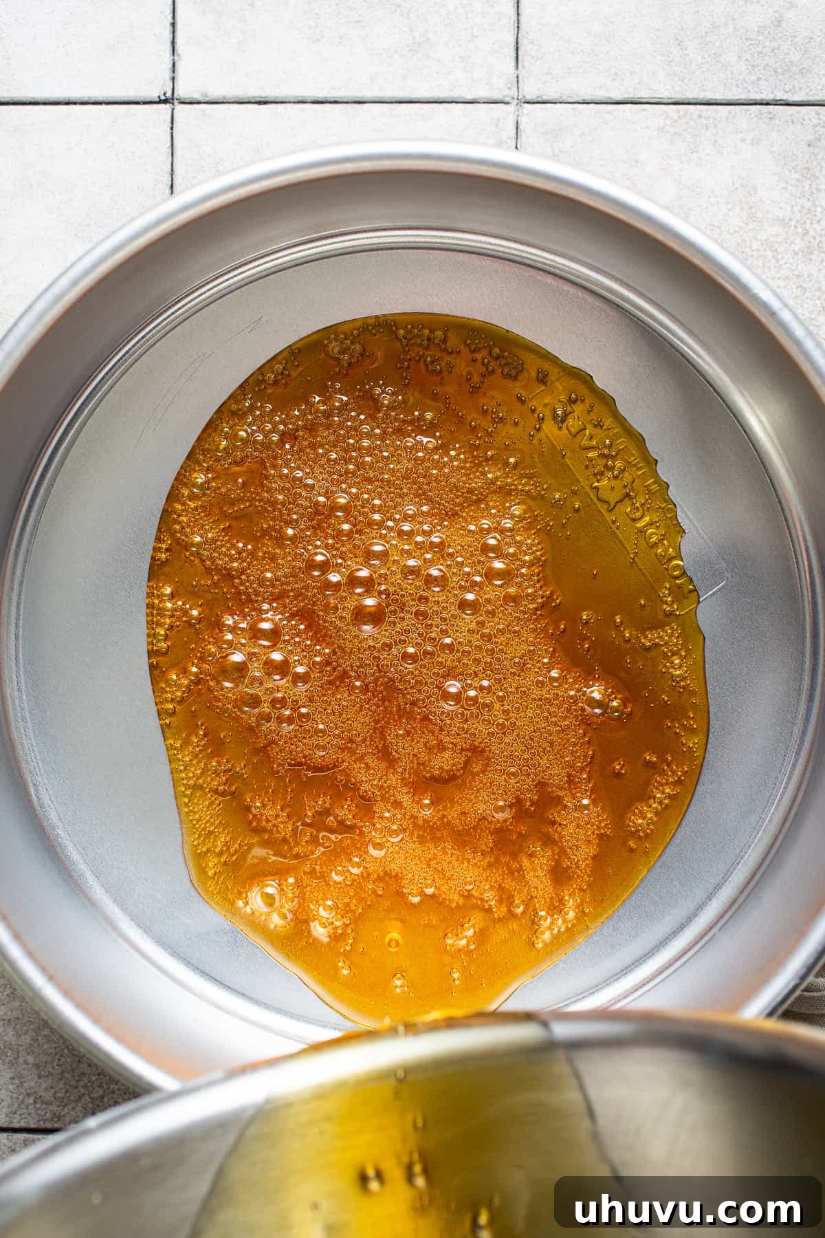 Rich Cheese Flan 4 Hot, golden-brown caramel being carefully poured into a round metal cake pan, evenly coating the bottom surface.