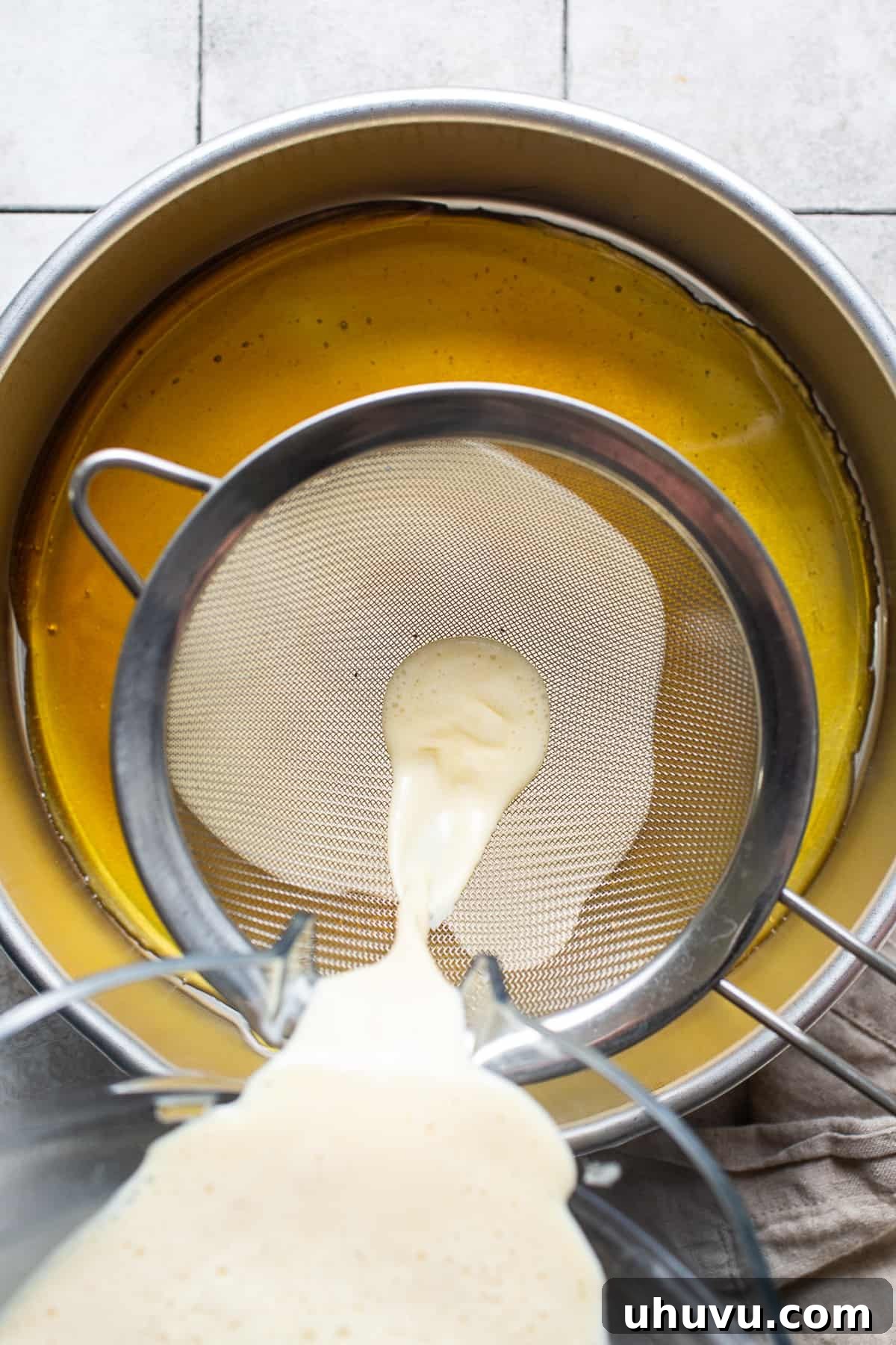 Rich Cheese Flan 5 The smooth, pale yellow flan mixture being poured through a fine mesh strainer into a round cake pan, which already contains hardened caramel at the bottom.