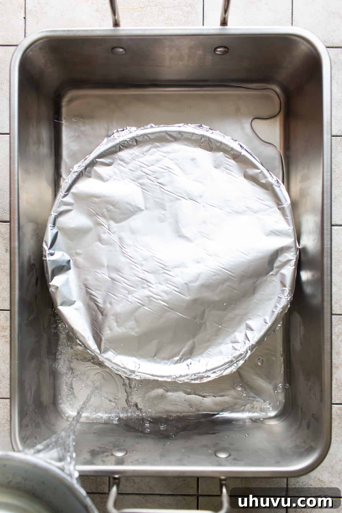 Rich Cheese Flan 6 A round cake pan, covered tightly with aluminum foil, resting inside a larger baking dish. Boiling water is being poured into the larger dish to create a bain-marie or water bath for baking.