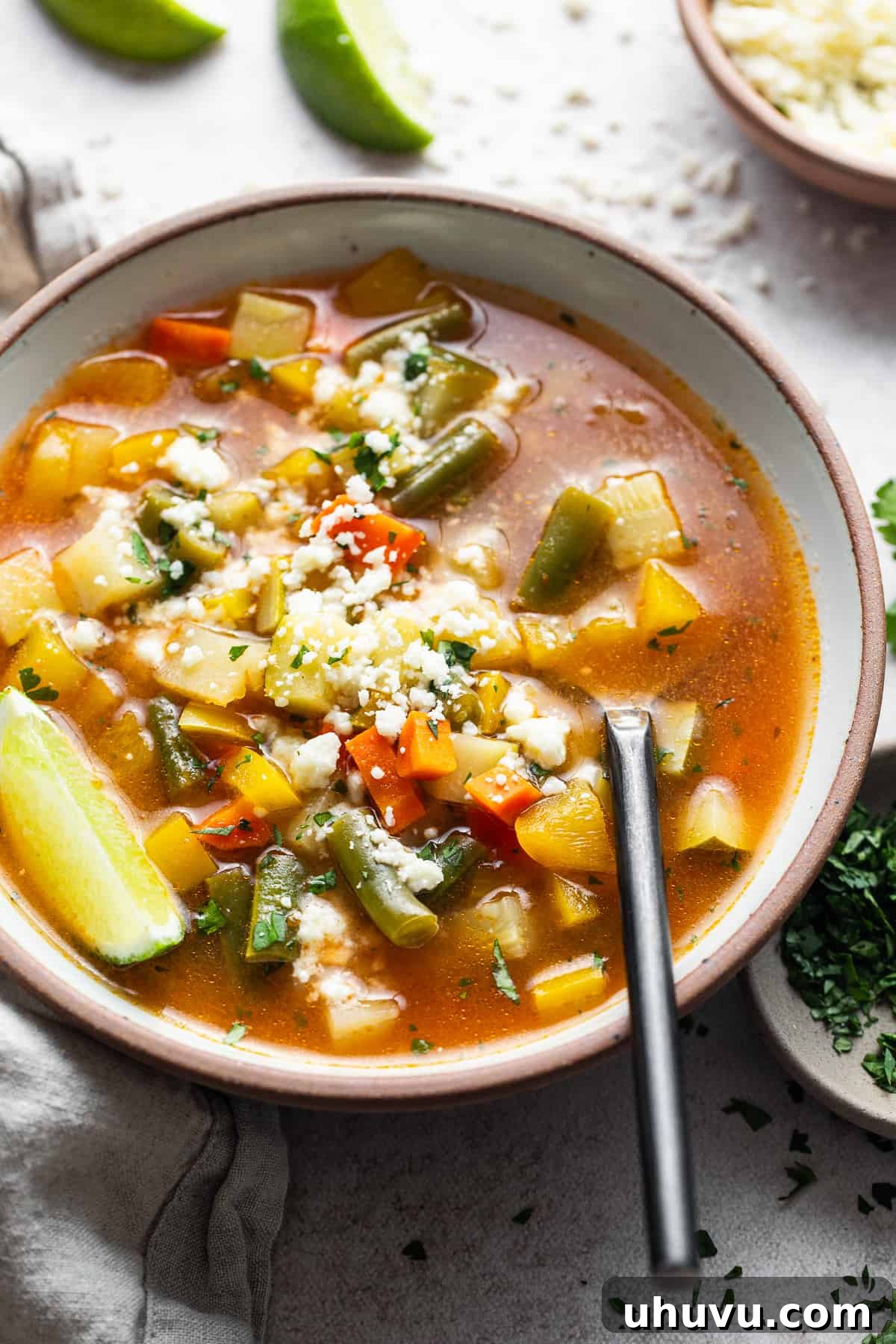 A warm bowl of Mexican vegetable soup garnished with lime wedges and sprinkled with cotija cheese, ready to be served.