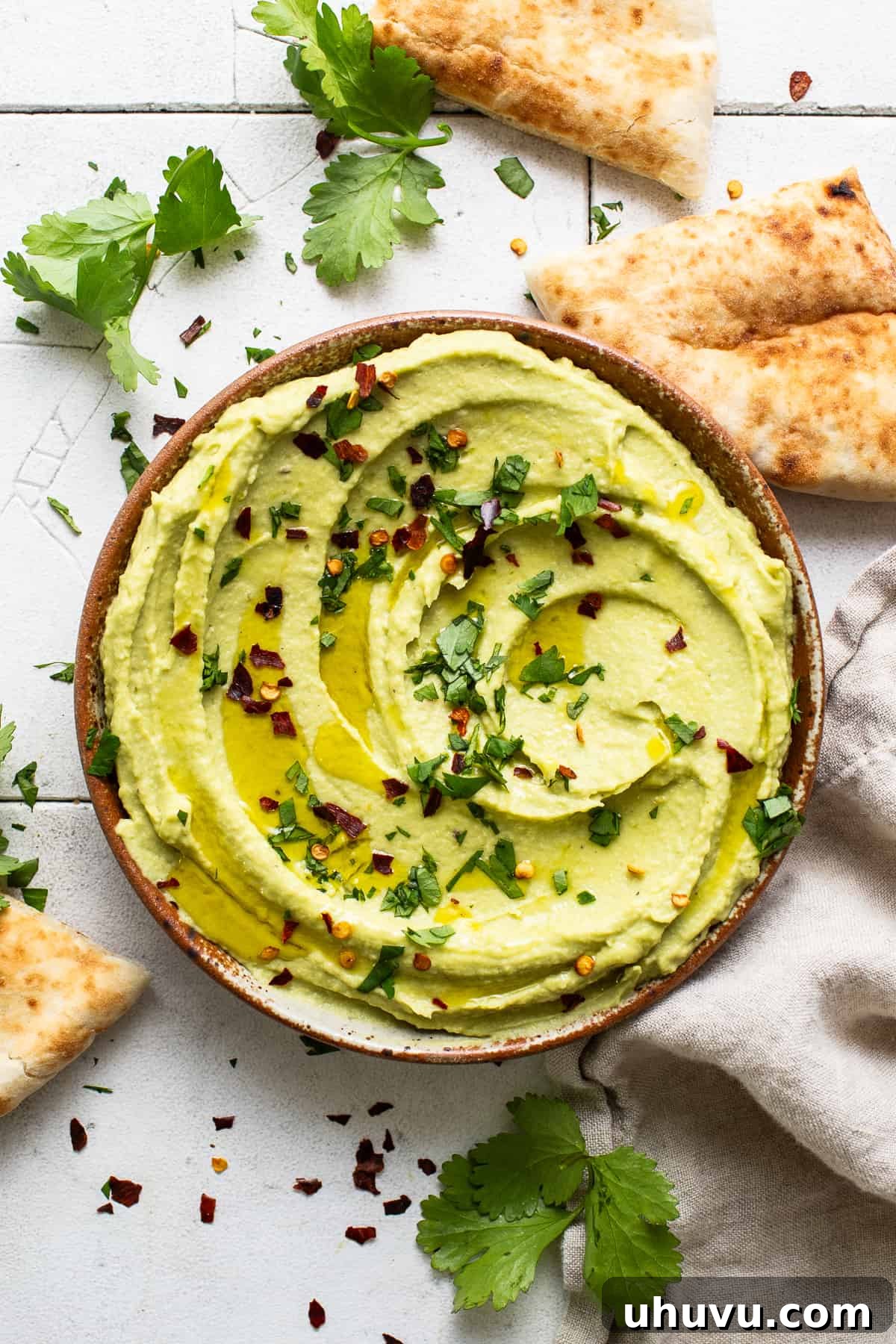Green Goddess Hummus 8 Avocado hummus in a stylish ceramic serving bowl drizzled with olive oil and garnished with chopped cilantro and red pepper flakes, placed on a light wooden surface.