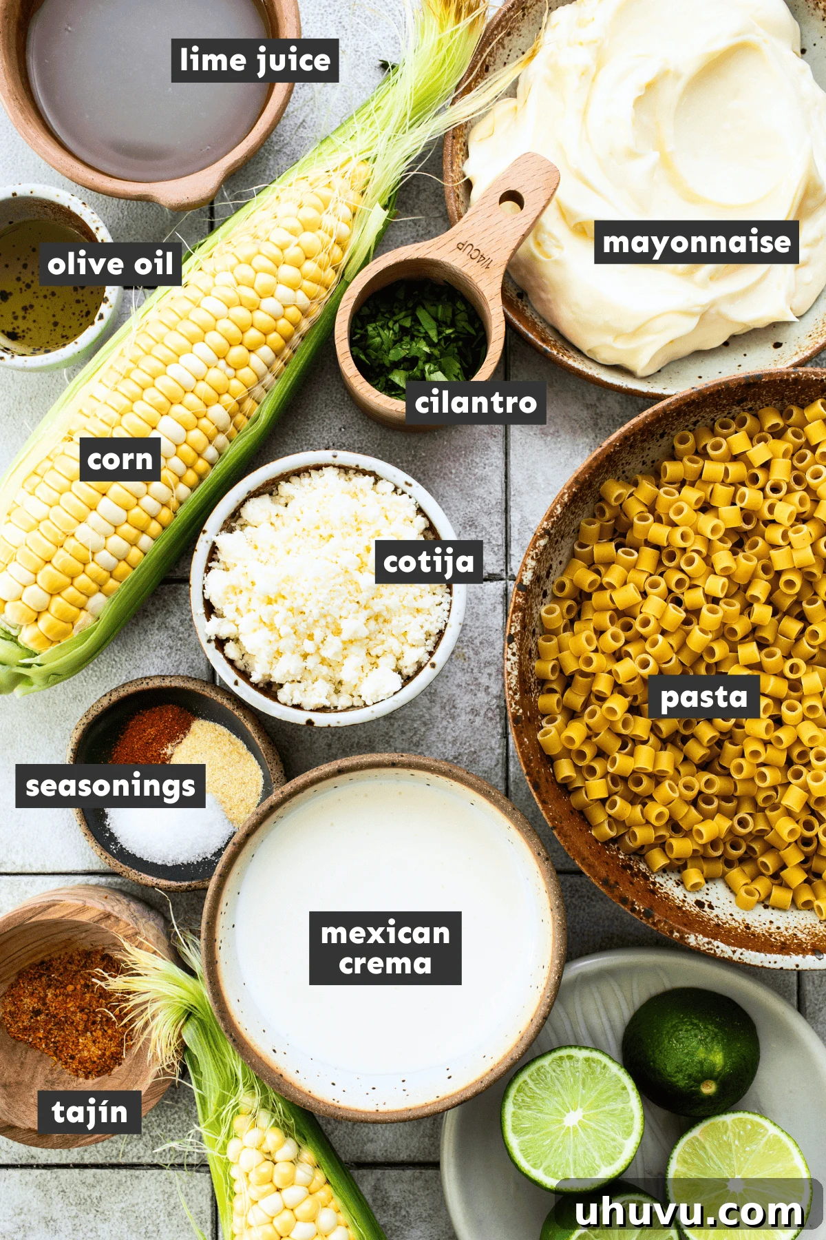 A flat lay photograph showcasing all the fresh ingredients for Mexican Street Corn Pasta Salad laid out on a table, including pasta, corn, lime, mayonnaise, cotija cheese, and cilantro.