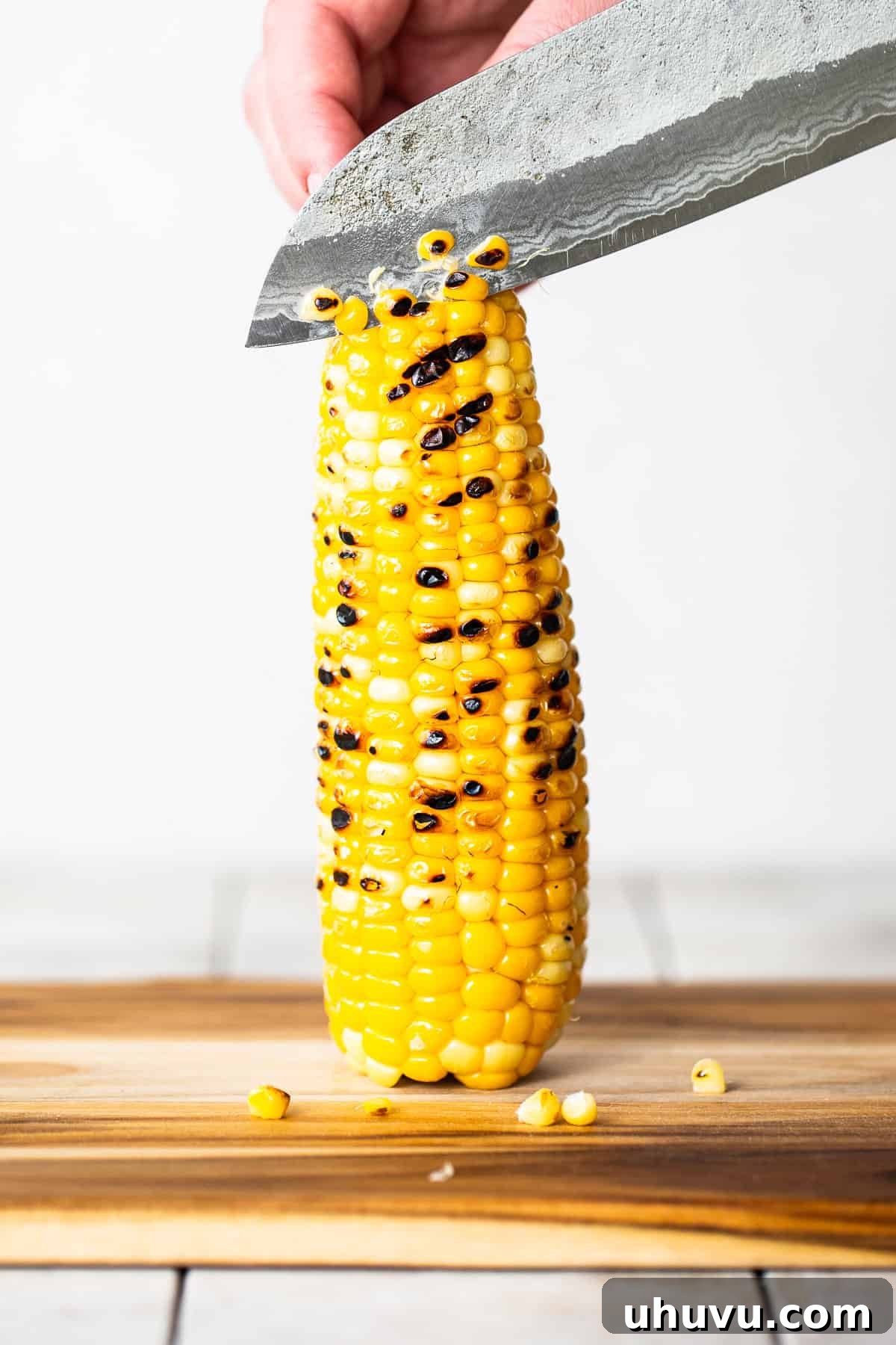 A chef's knife carefully slicing vibrant yellow corn kernels off a fire-roasted cob into a bowl, preparing them for the pasta salad.