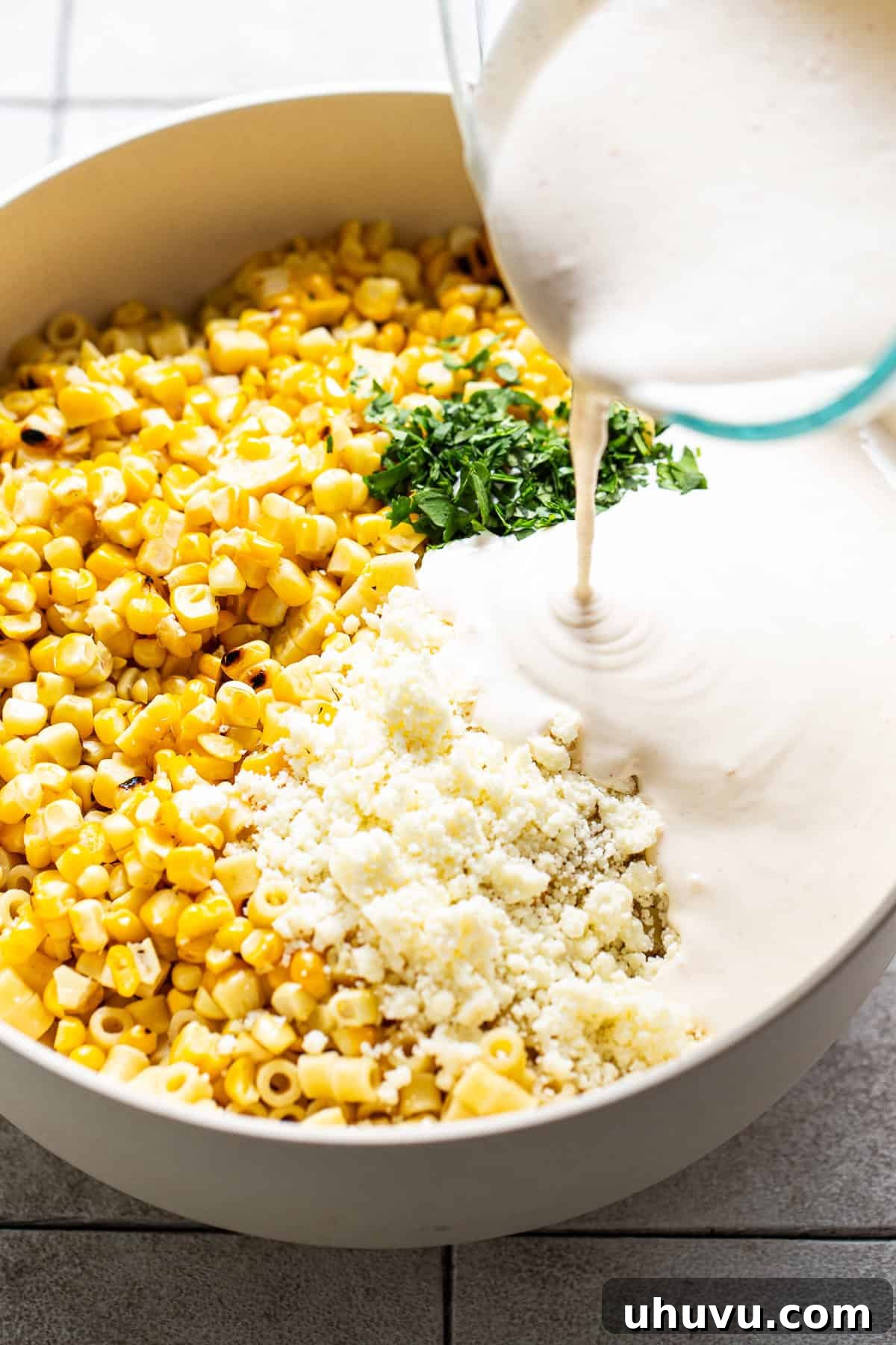 Various ingredients for Mexican Street Corn Pasta Salad, including cooked pasta, corn, cilantro, and cotija cheese, being combined in a large mixing bowl with the creamy dressing.