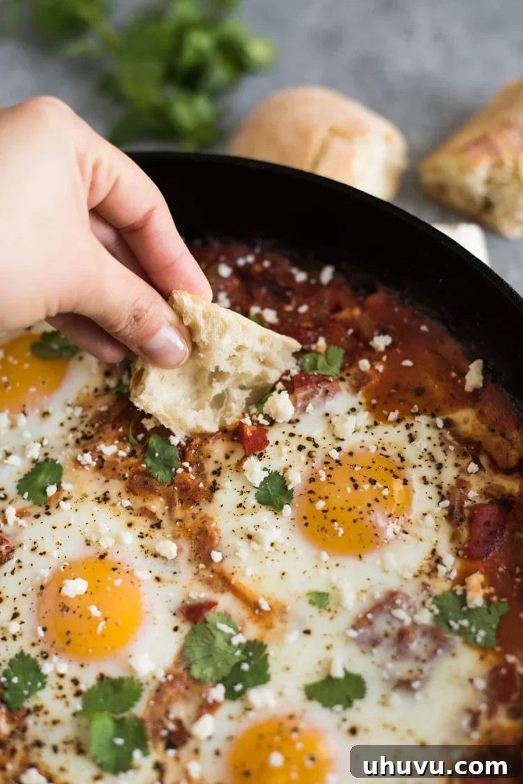 Crusty bread being generously dipped into a rich, steaming Shakshuka, highlighting the perfect runny yolk and the vibrant tomato sauce infused with feta cheese.