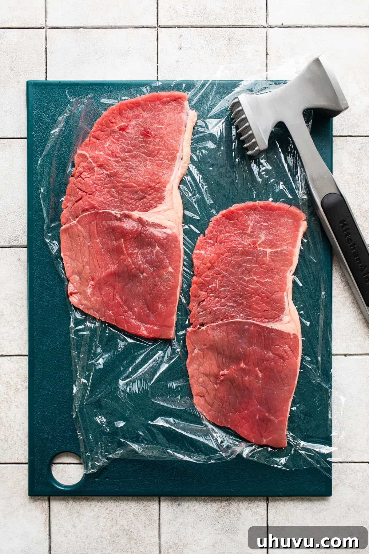 Thinly sliced beef steak after being pounded with a meat mallet on a cutting board lined with plastic wrap, ready for breading.
