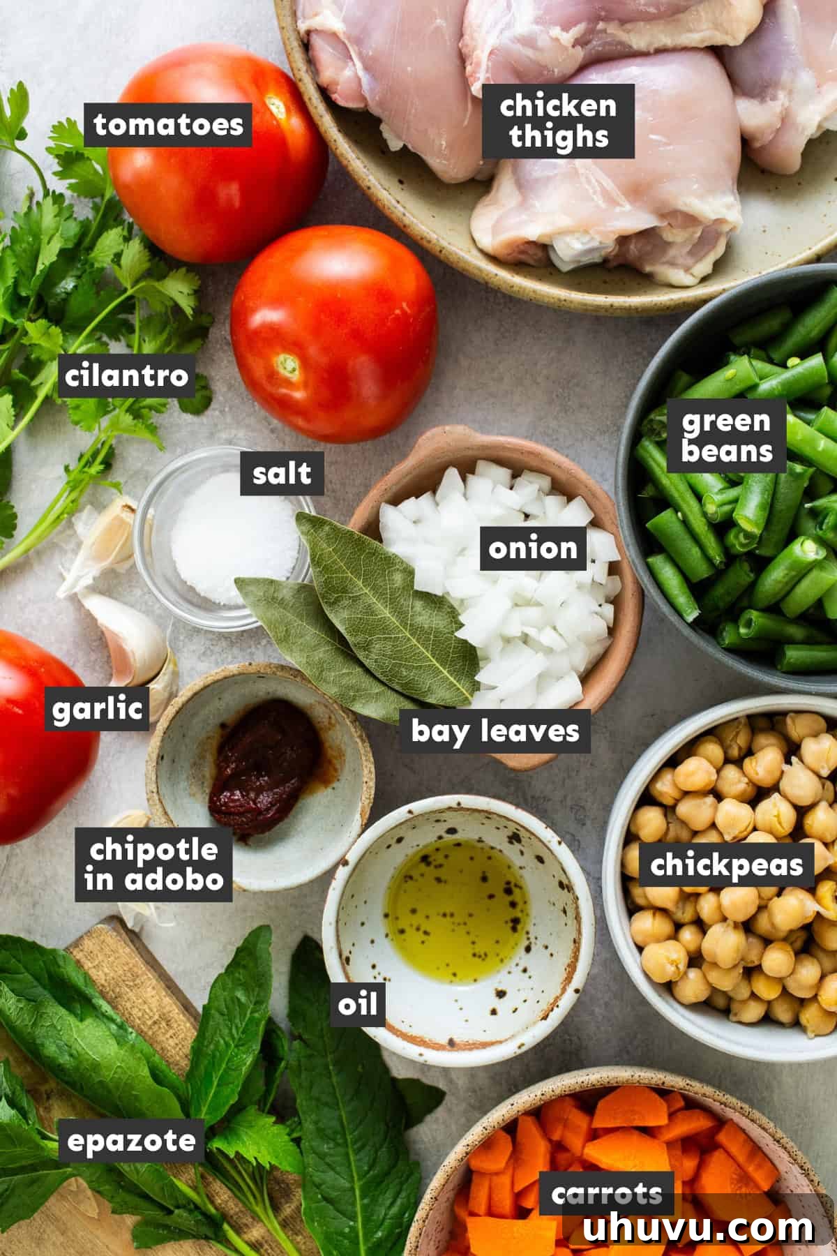 A flat lay photograph showcasing all the fresh ingredients for Caldo Tlalpeño laid out on a wooden table, including chicken, tomatoes, onions, garlic, carrots, green beans, chickpeas, and herbs.