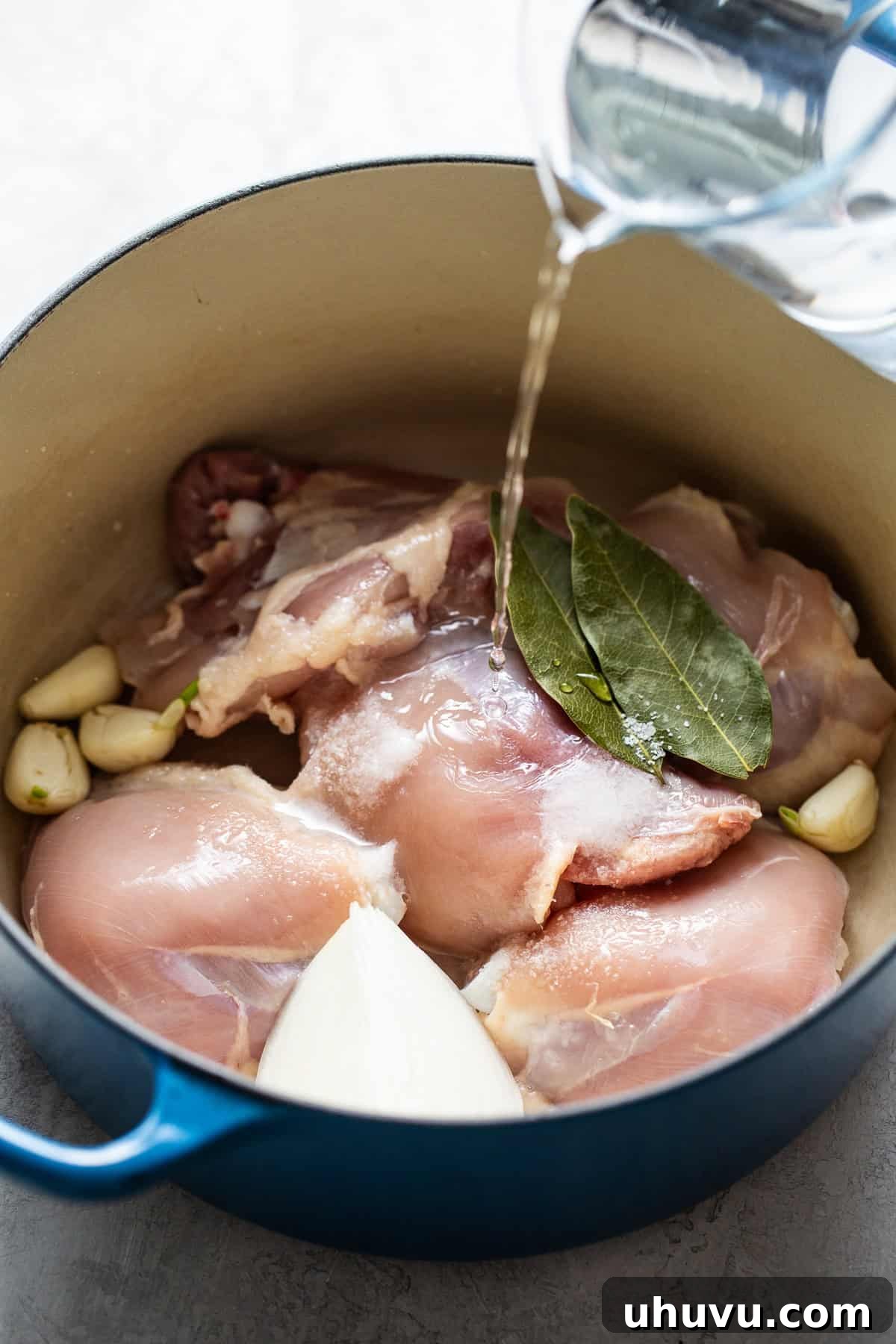 Pouring water into a large pot containing bone-in chicken thighs, roughly chopped white onion, garlic cloves, and bay leaves, initiating the process of making Mexican chicken broth.