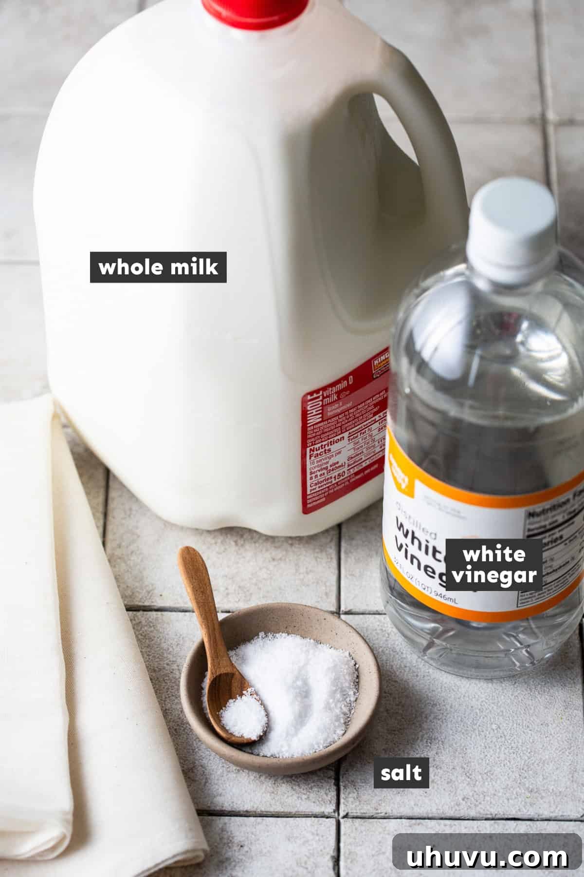 Make Your Own Queso Fresco 3 A simple array of ingredients for making queso fresco, including a gallon of whole milk, a bottle of distilled white vinegar, and a container of kosher salt, all neatly arranged on a rustic wooden table.