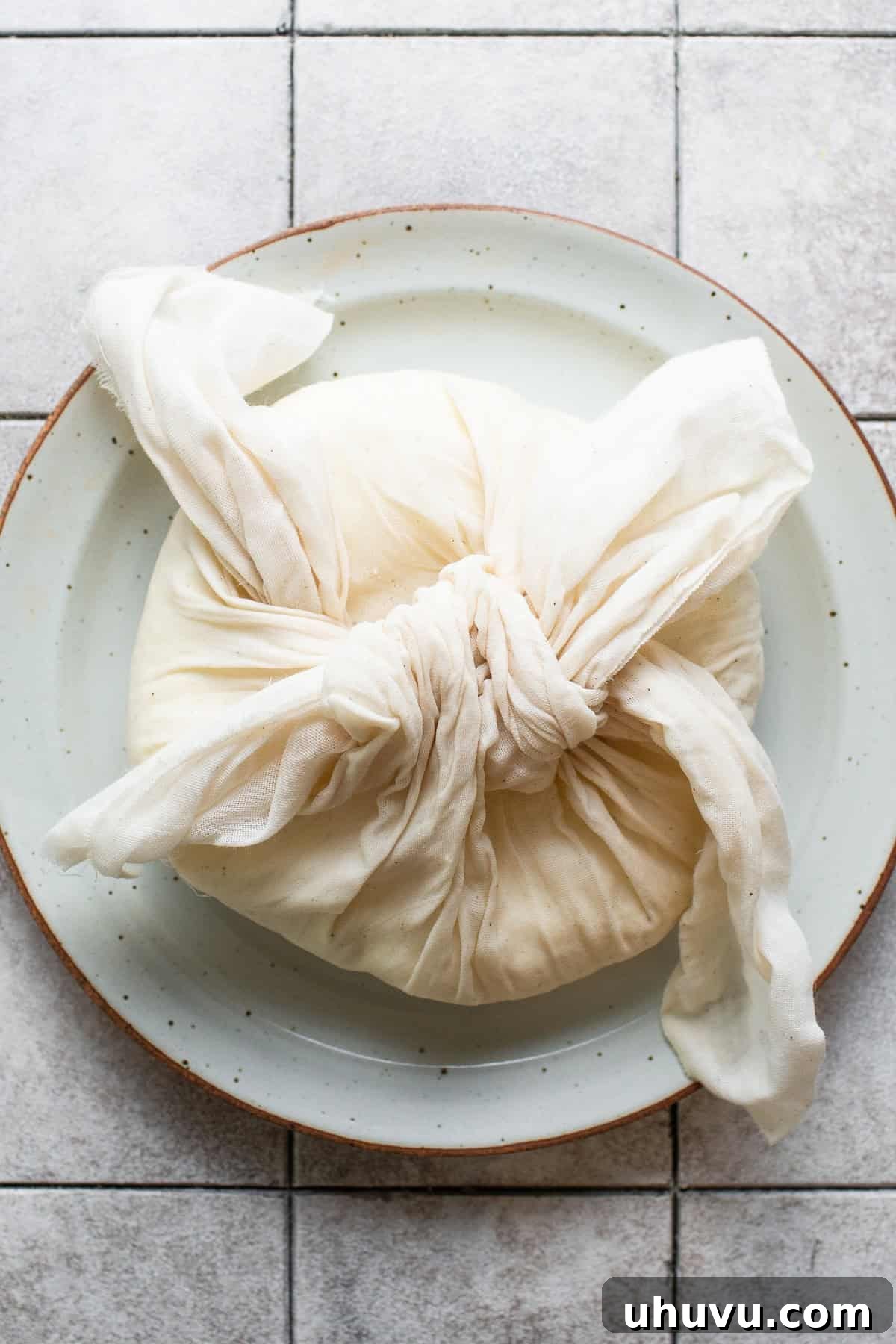 Make Your Own Queso Fresco 7 A bundle of queso fresco wrapped and tied in cheesecloth, weighted down by a heavy can in a colander, illustrating the final pressing stage.