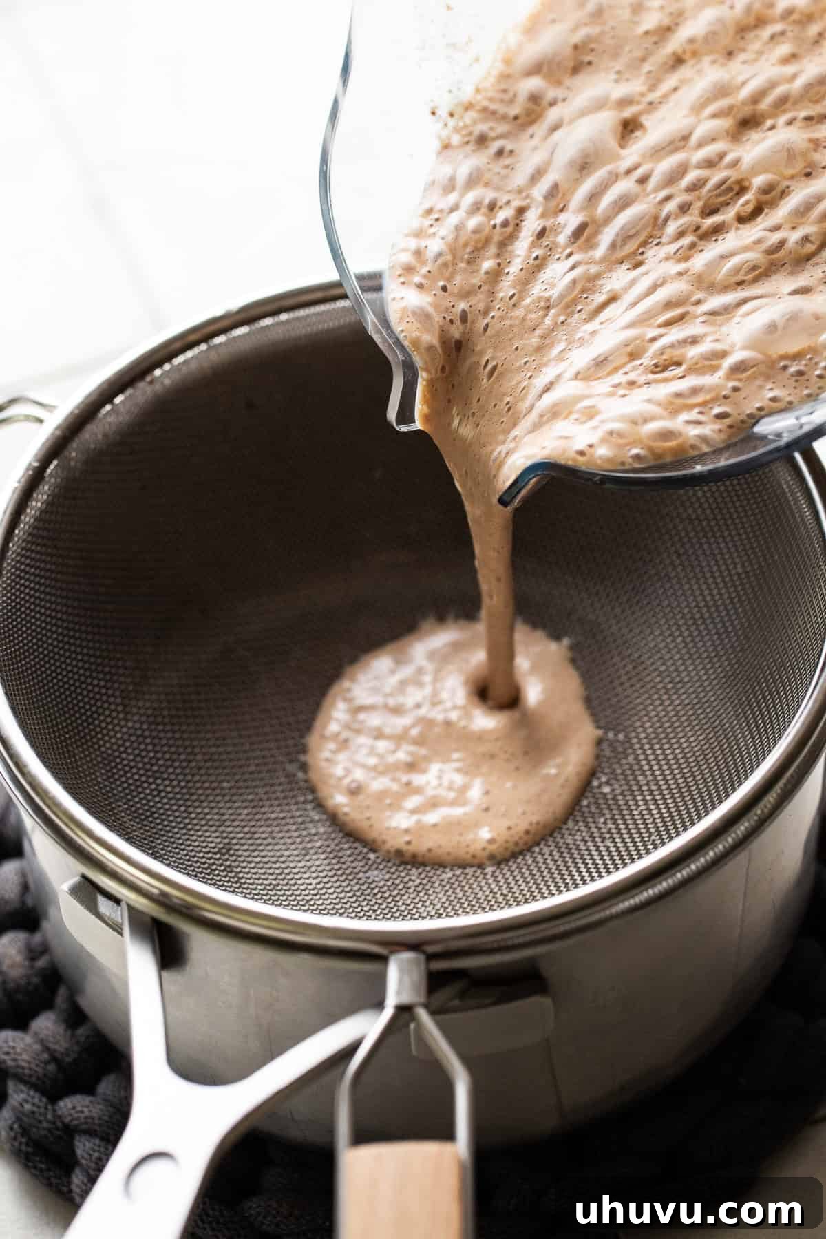 Cozy Walnut Brew 7 The smooth, blended atole de nuez mixture being carefully poured through a fine-mesh strainer into a pot on the stove, ensuring a silky texture.