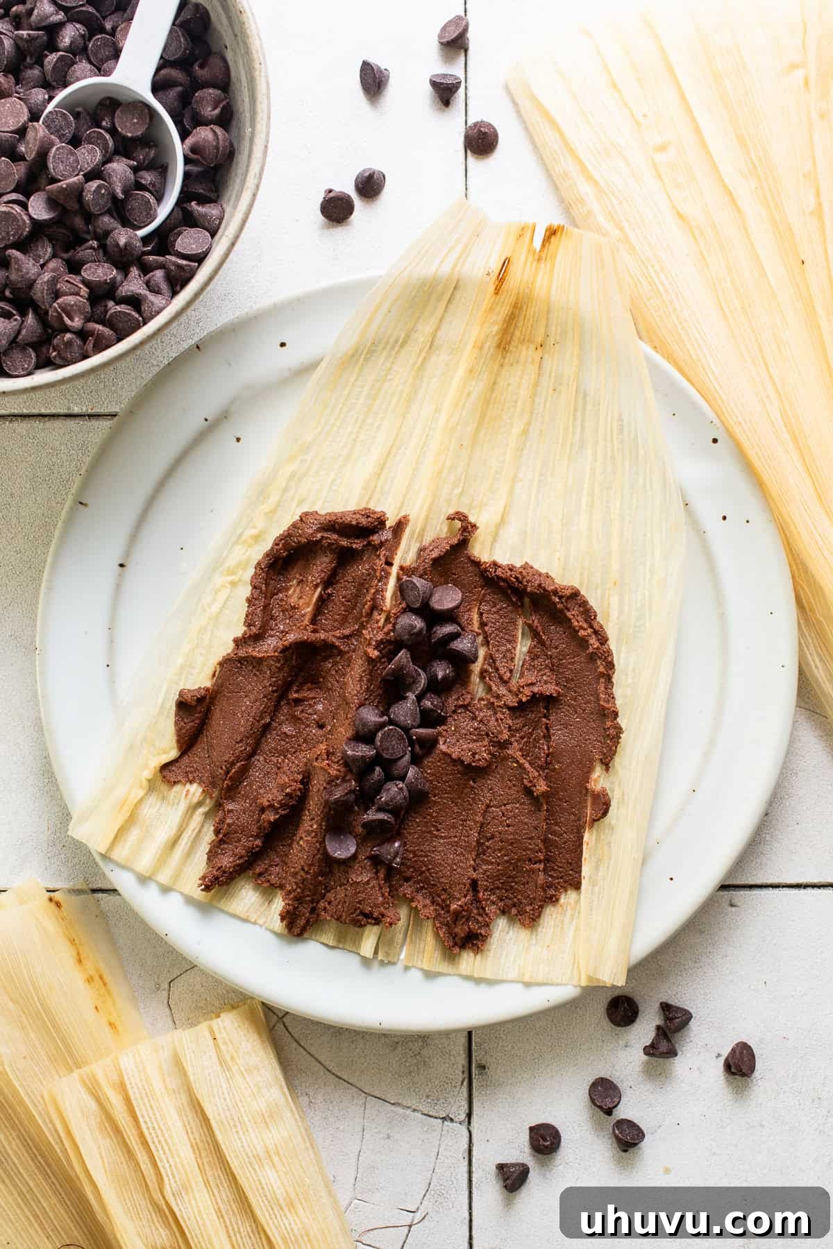 Chocolate masa dough spread generously on a softened corn husk, with a spoonful of semi-sweet chocolate chips nestled in the center, ready for folding.
