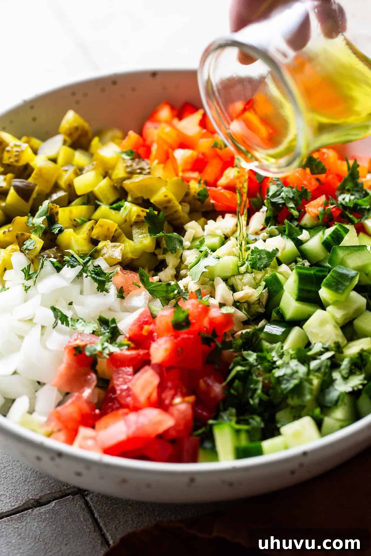 Briny Salsa 5 Pickle brine being poured into a large mixing bowl filled with finely diced pickles, cucumbers, tomatoes, and other fresh ingredients for pickle de gallo.