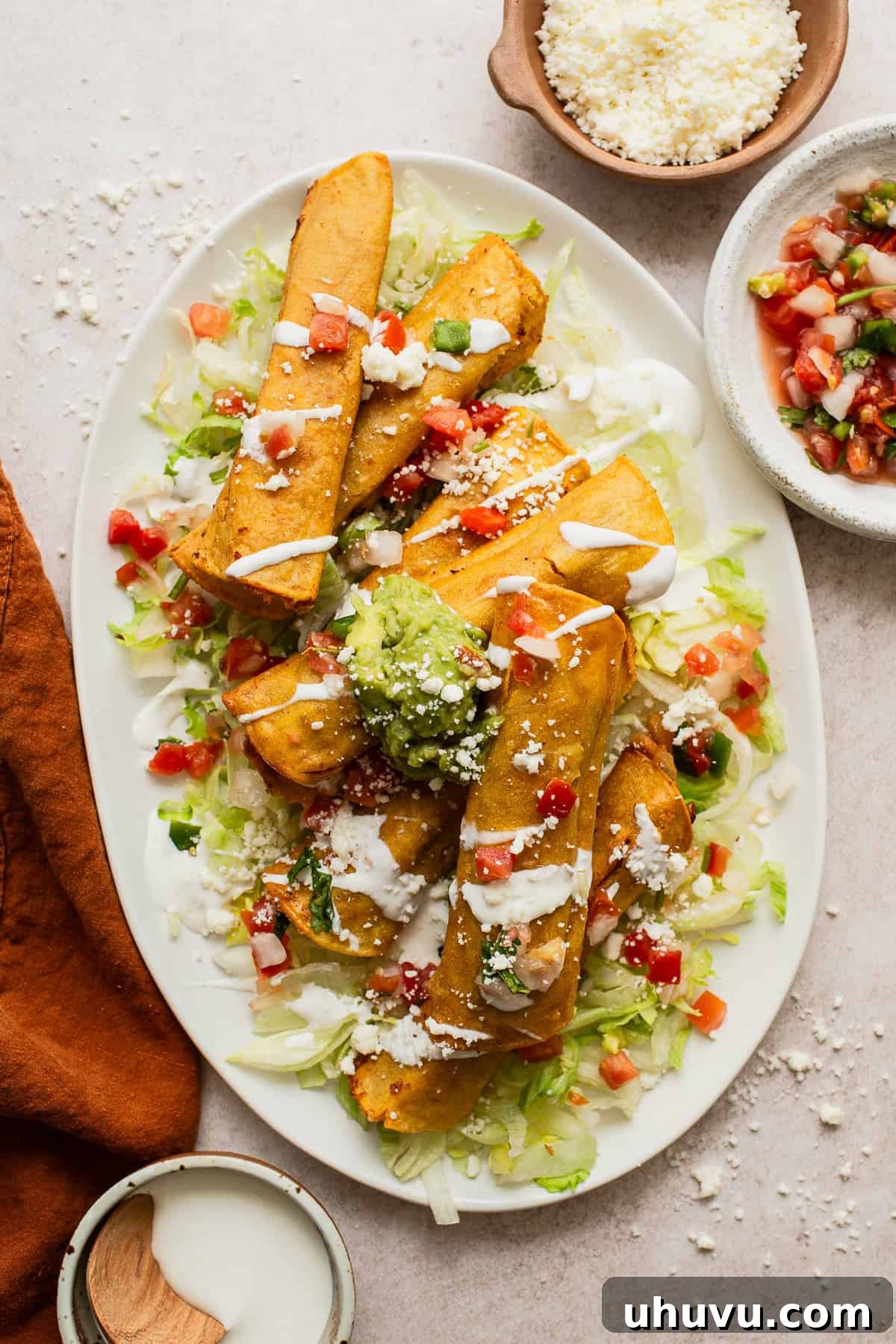 Crispy bean and cheese taquitos on a large plate, garnished with fresh shredded lettuce, a drizzle of Mexican crema, diced tomatoes, creamy guacamole, and crumbled cotija cheese. A perfect, vibrant serving.
