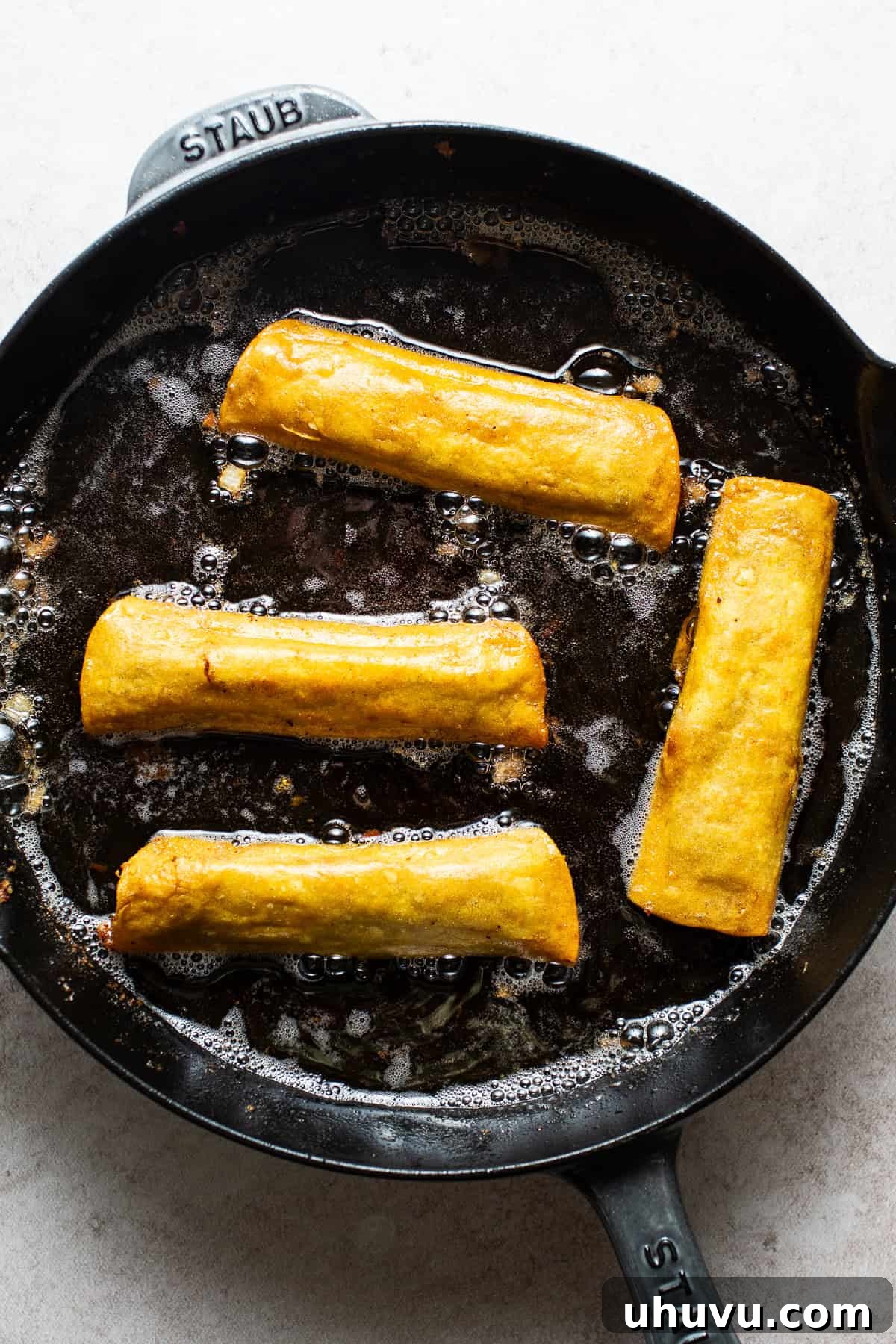 Several bean and cheese taquitos sizzling and turning golden brown in hot oil in a skillet.