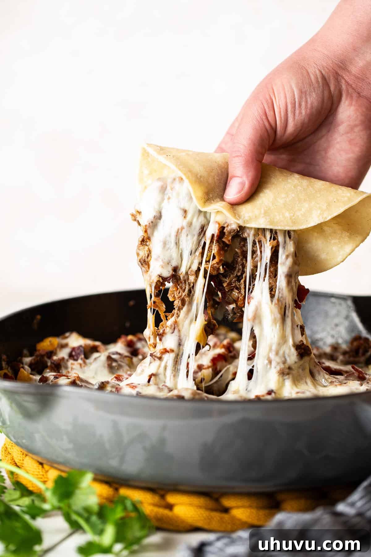 A warm corn tortilla being used to scoop a generous portion of cheesy Tacos de Alambre filling directly from the skillet, demonstrating the family-style serving.