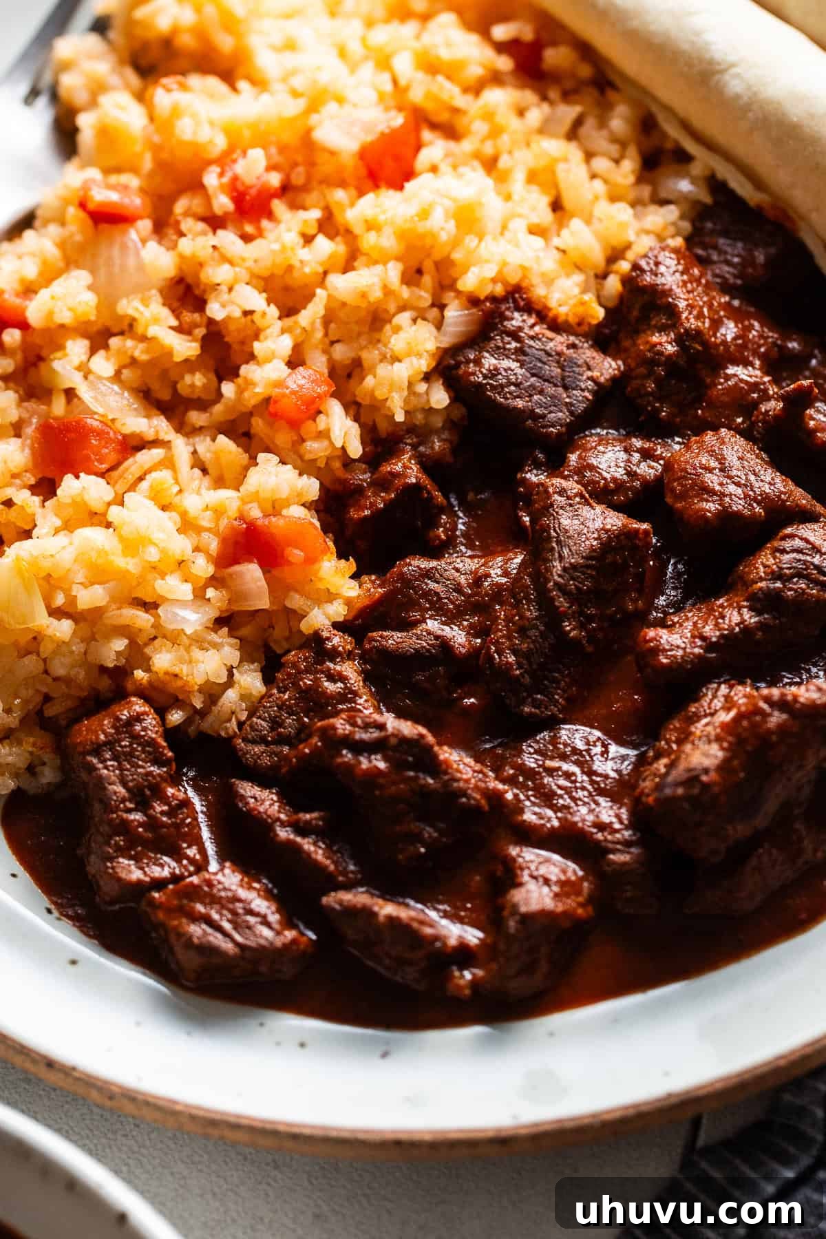 Traditional chile colorado made from tender pieces of beef in a red chile sauce on a plate next to red Mexican rice.
