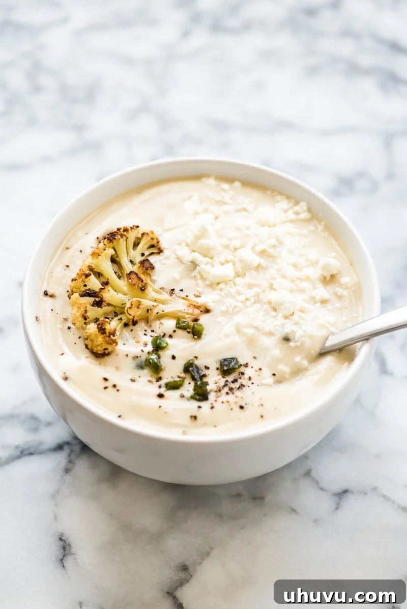Overhead shot of Roasted Cauliflower Soup in a cozy bowl, garnished with a swirl of plant-based cream, fried jalapeños, and fresh herbs, perfect for a warming meal.