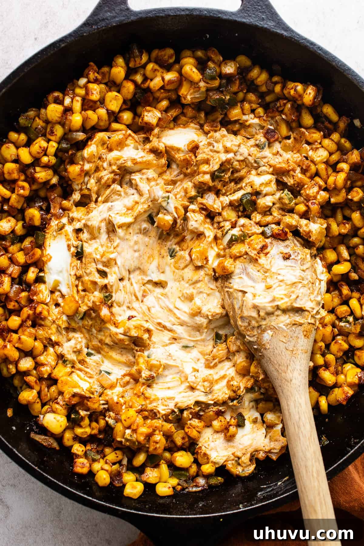 Elote Fiesta Dip 5 Softened cream cheese being stirred into a cast iron skillet with corn, cooked onions, and jalapeños, creating a creamy texture.