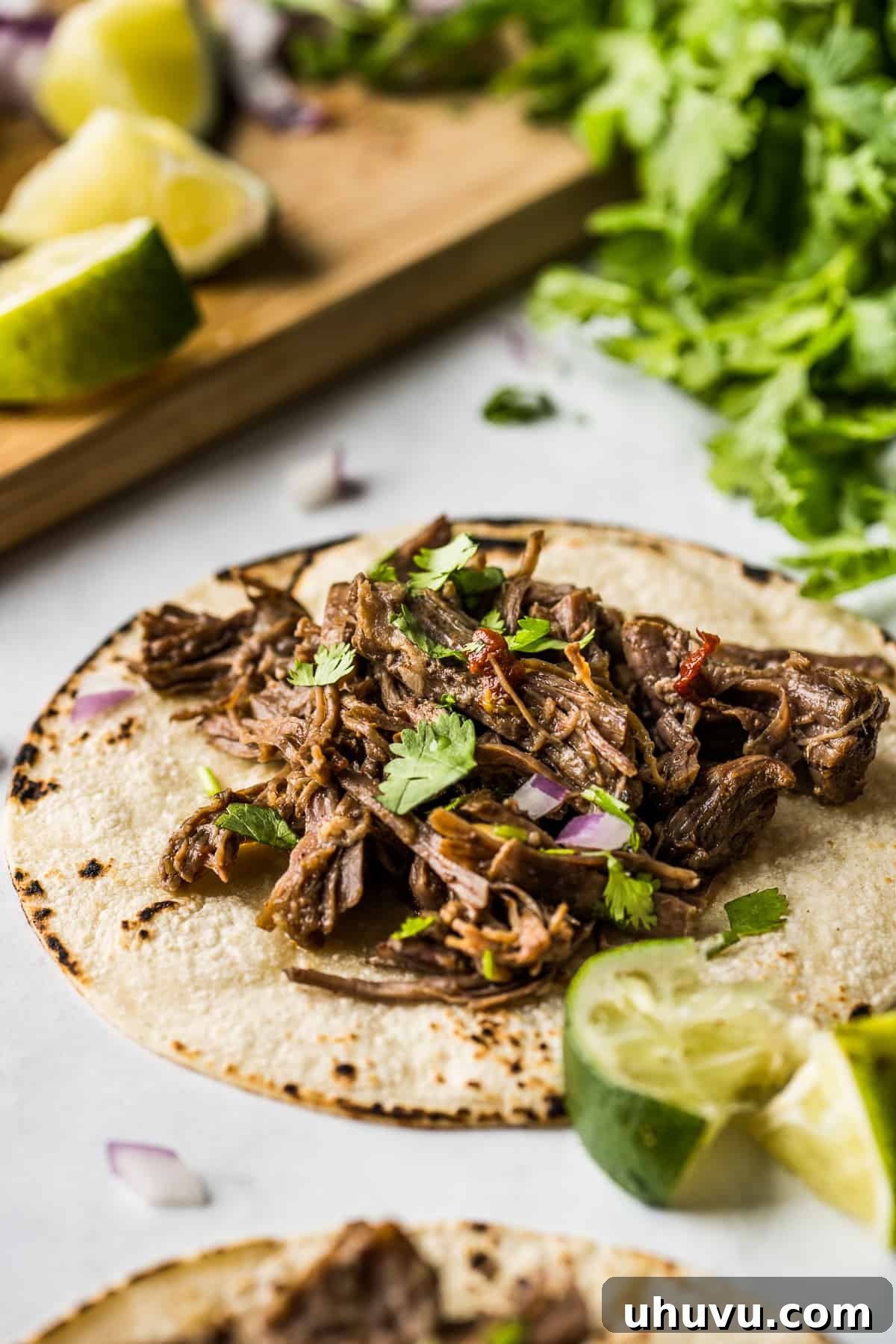 Melt-in-Your-Mouth Barbacoa 12 A close-up of a corn tortilla overflowing with tender barbacoa meat, garnished with fresh lime juice, finely diced red onions, and vibrant cilantro.