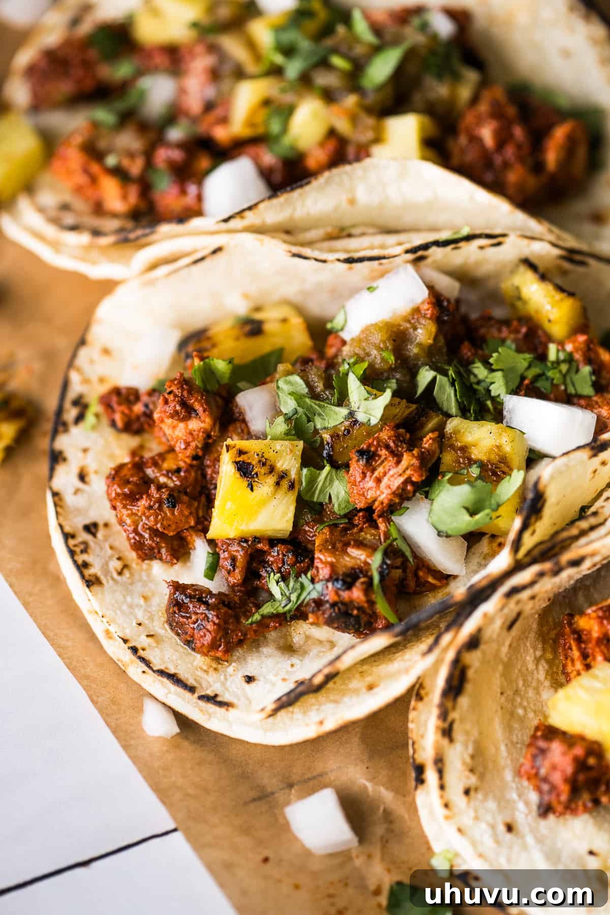 Fully assembled Tacos al Pastor in warm corn tortillas, generously topped with grilled pineapple, fresh cilantro, diced white onion, and a drizzle of vibrant salsa verde.
