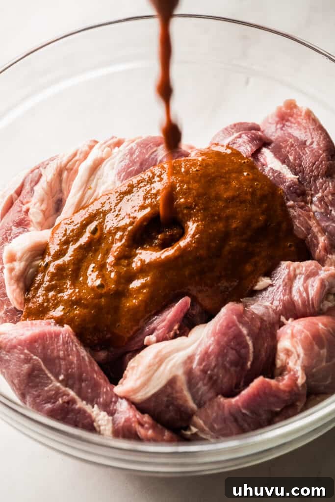 Vibrant red marinade being poured over thin slices of pork shoulder in a glass bowl.