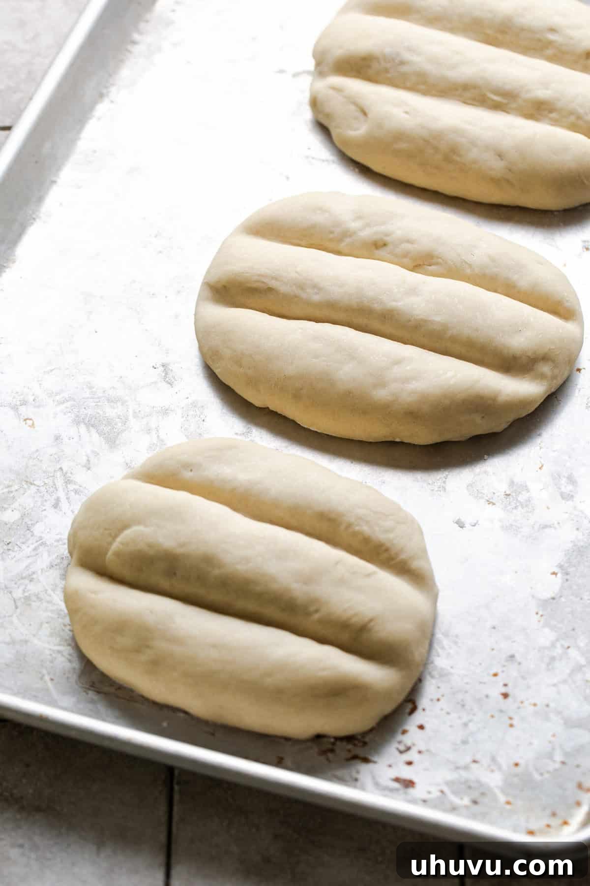 Telera bread rolls on a baking sheet, covered with a towel for their second rise.